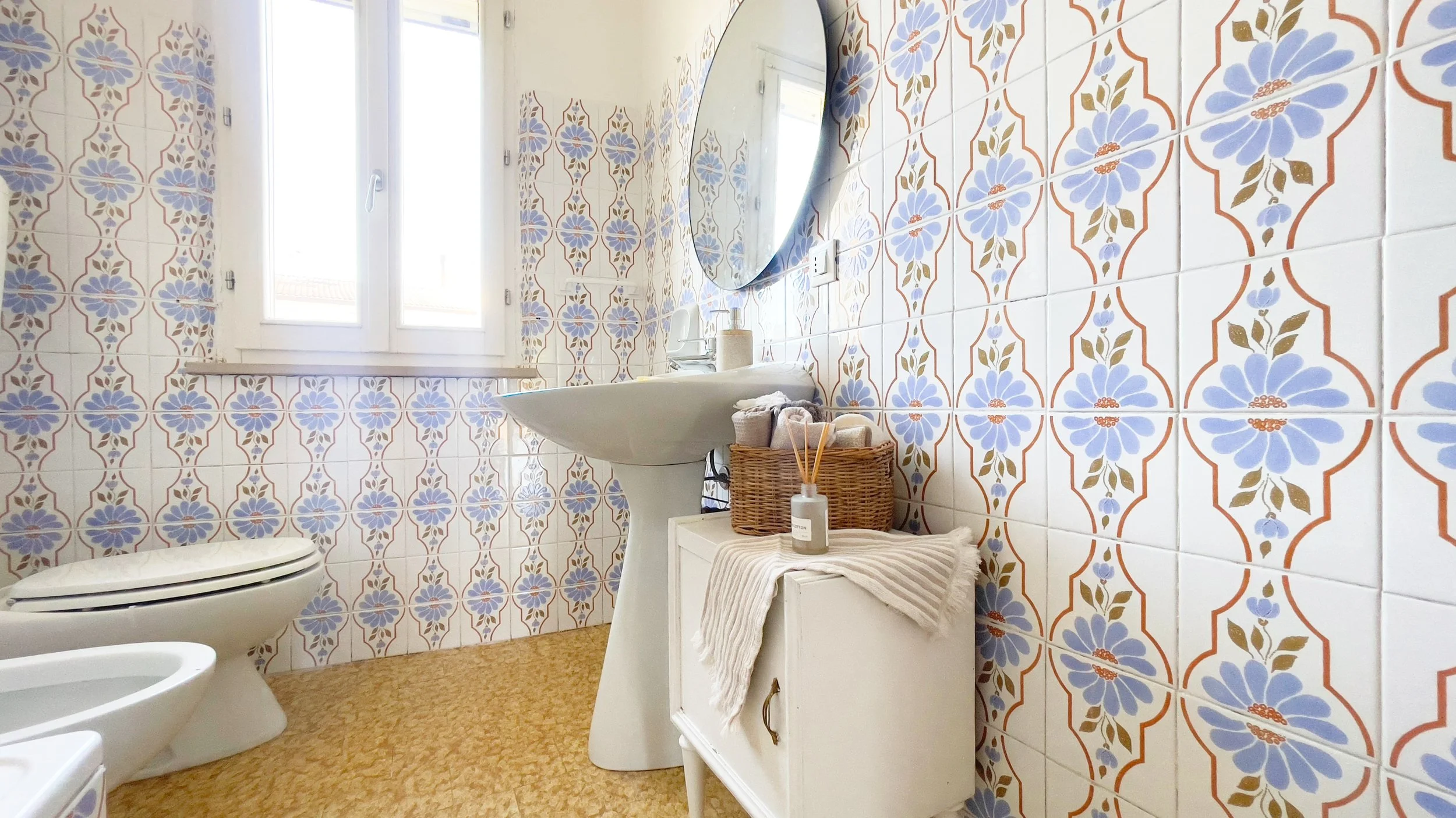 Bagno con piastrelle floreali colorate, lavabo bianco, specchio rotondo, finestra luminosa, mobiletto con cesto di asciugamani e diffusore di profumo.