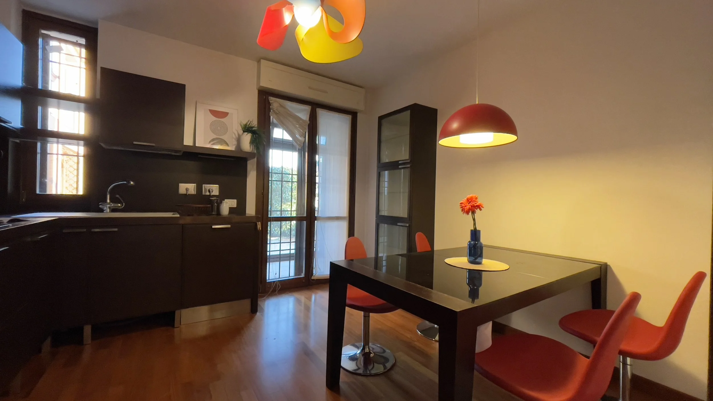 Cucina moderna con mobili scuri, tavolo da pranzo nero e sedie rosse, lampade colorate e finestra luminosa.