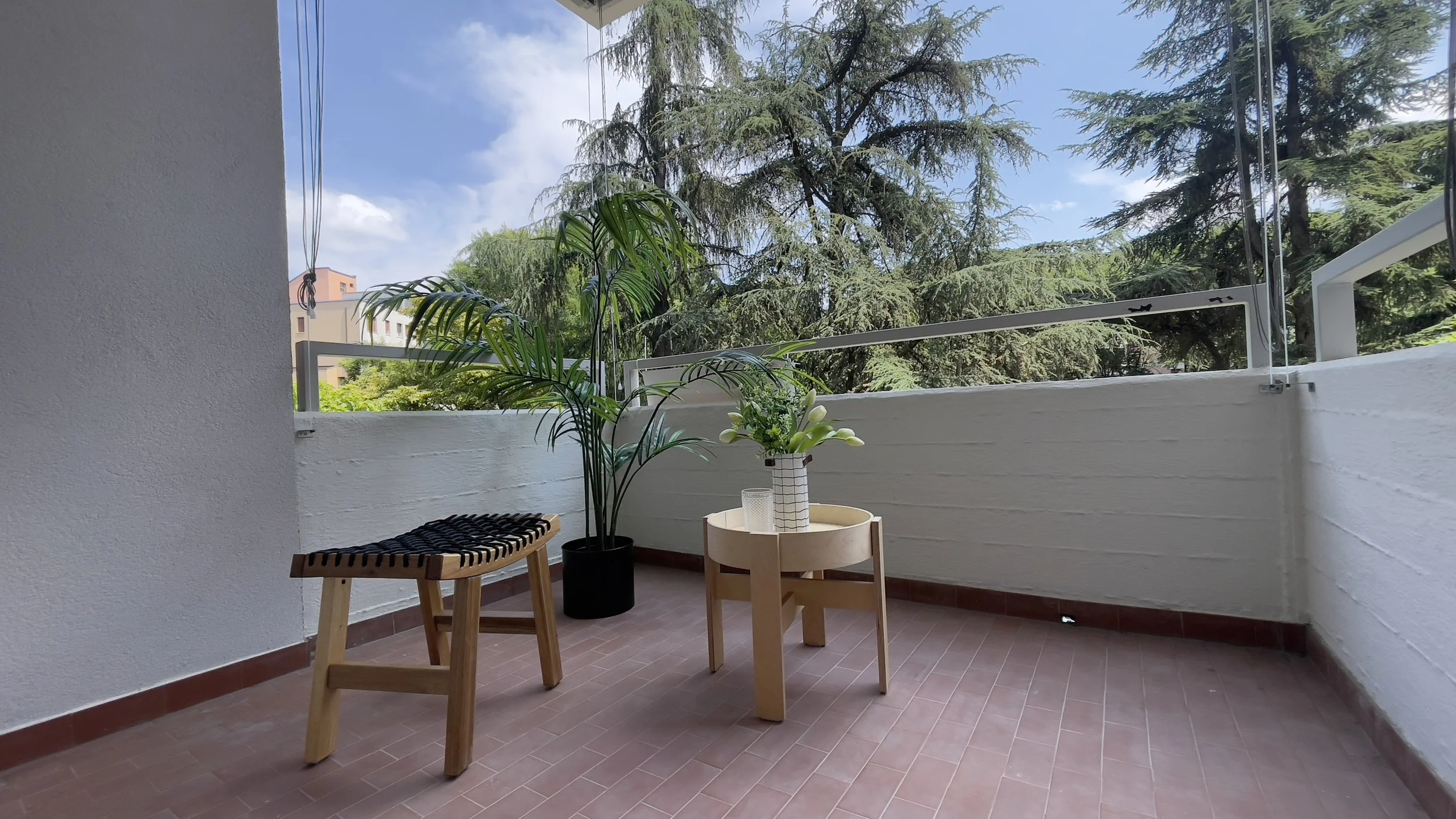 Terrazzo arredato con piante, tavolino, sgabello, cielo e alberi sullo sfondo.