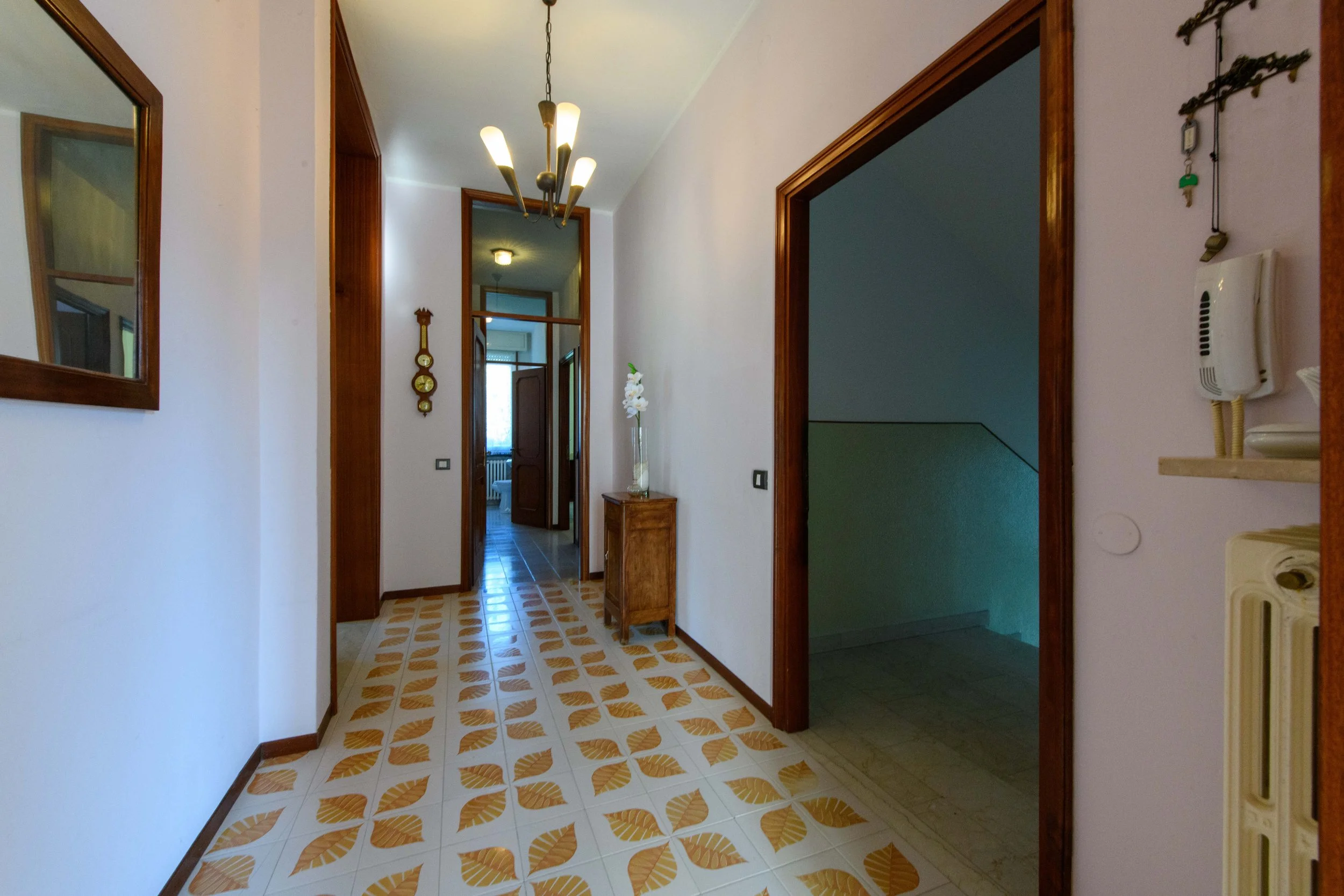 Un corridoio interno con pavimento decorato, pareti chiare, un lampadario moderno, una specchiera sulla sinistra e un piccolo mobile con un vaso di fiori sulla destra. Verso il fondo si vedono altre stanze con porte aperte.