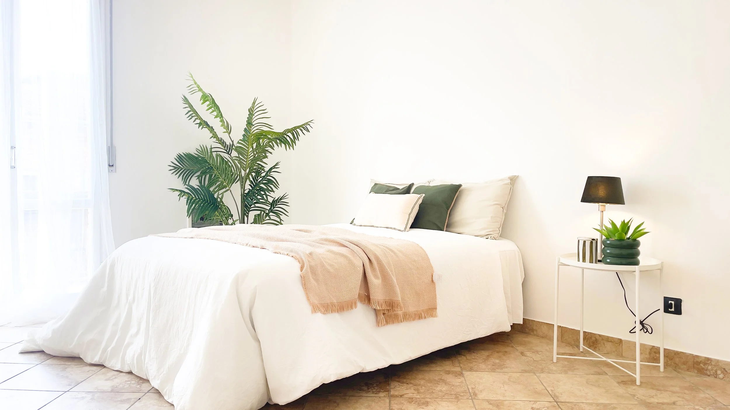 Camera da letto luminosa con letto matrimoniale, cuscini e plaid, pianta verde accanto e tavolino bianco con lampada e pianta in vaso.