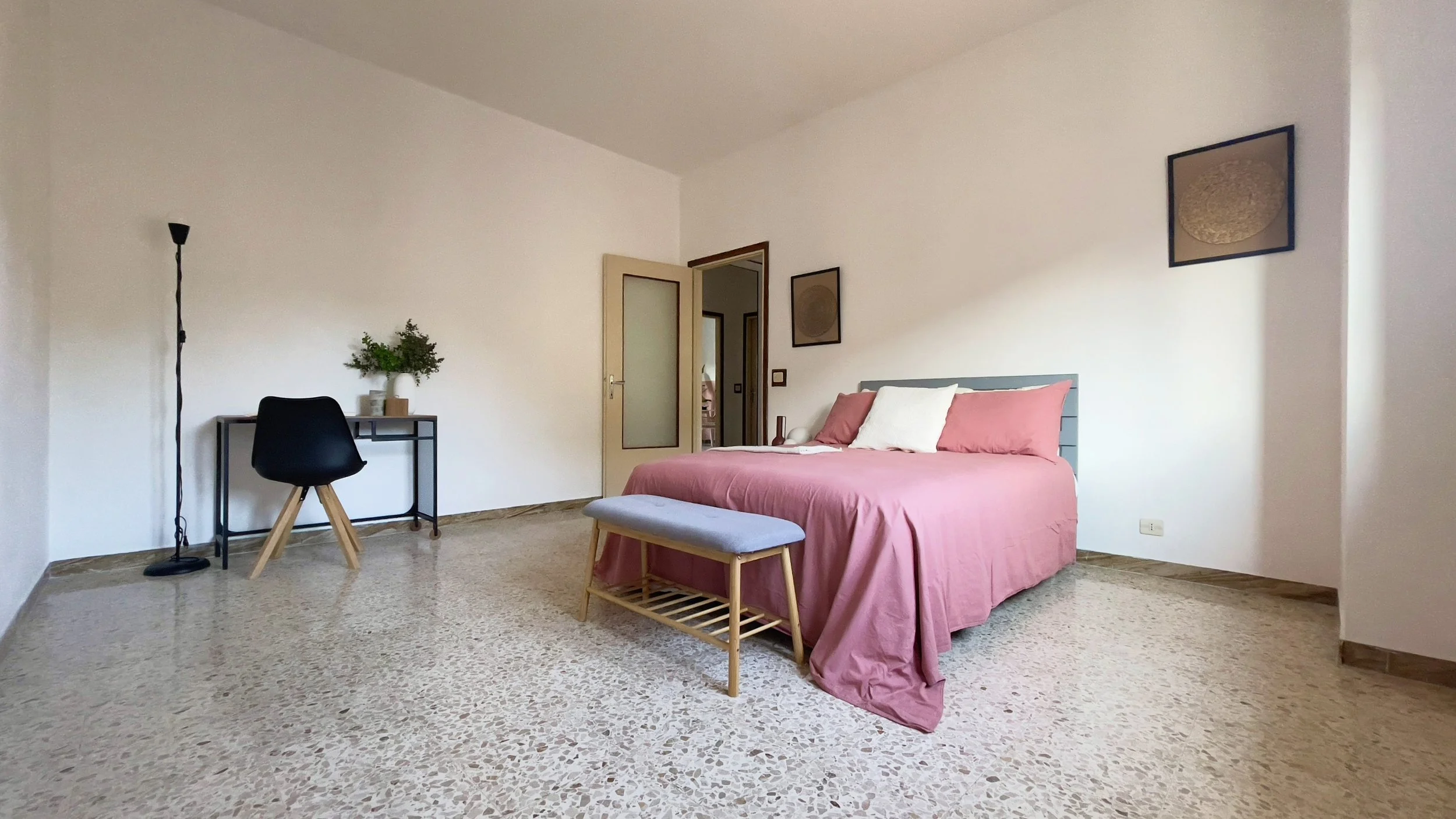 Camera da letto con letto matrimoniale con copriletto rosa, due cuscini, panca ai piedi del letto, scrivania nera con sedia e pianta decorativa, lampada da terra, pavimento in granito e due quadri sulle pareti.