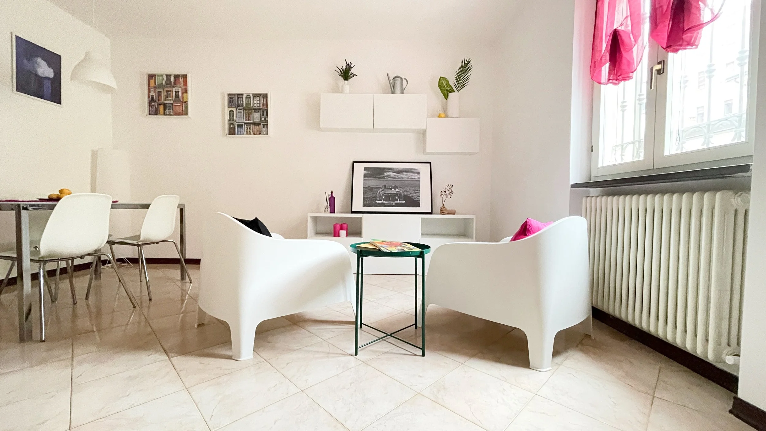 Interno di un soggiorno moderno con due poltrone bianche, un piccolo tavolino verde, una finestra con tende rosa chiaro e un tavolo da pranzo con sedie bianche. Il pavimento è piastrellato e ci sono opere d'arte appese alle pareti bianche. Piante e u