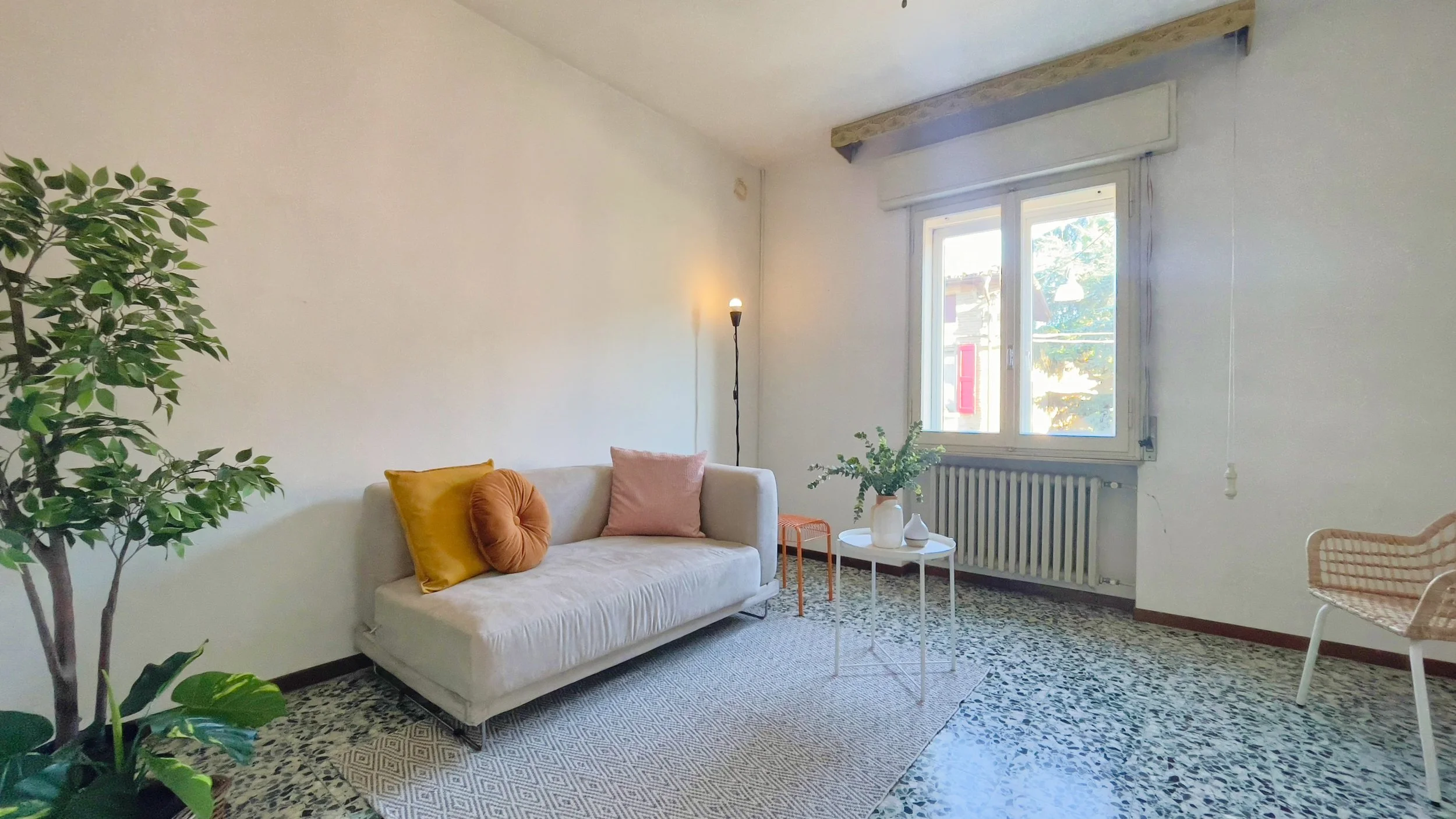 Soggiorno luminoso con divano beige, cuscini colorati, un tavolino con decorazioni floreali, pianta verde e pavimento in marmo.