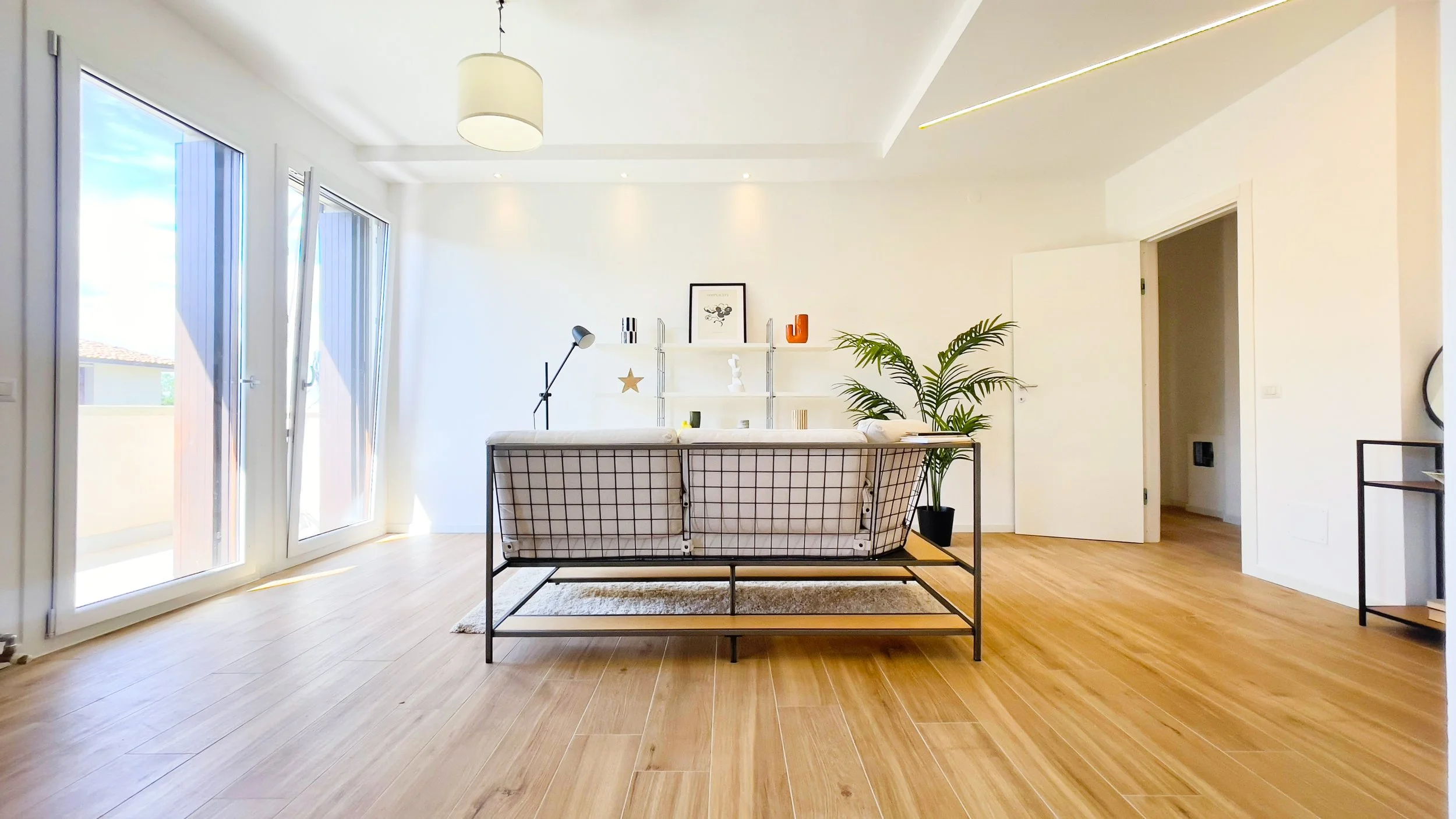 Soggiorno moderno con divano, lampada da terra, mensole e pianta decorativa, pavimento in legno e porta a vetri aperta.