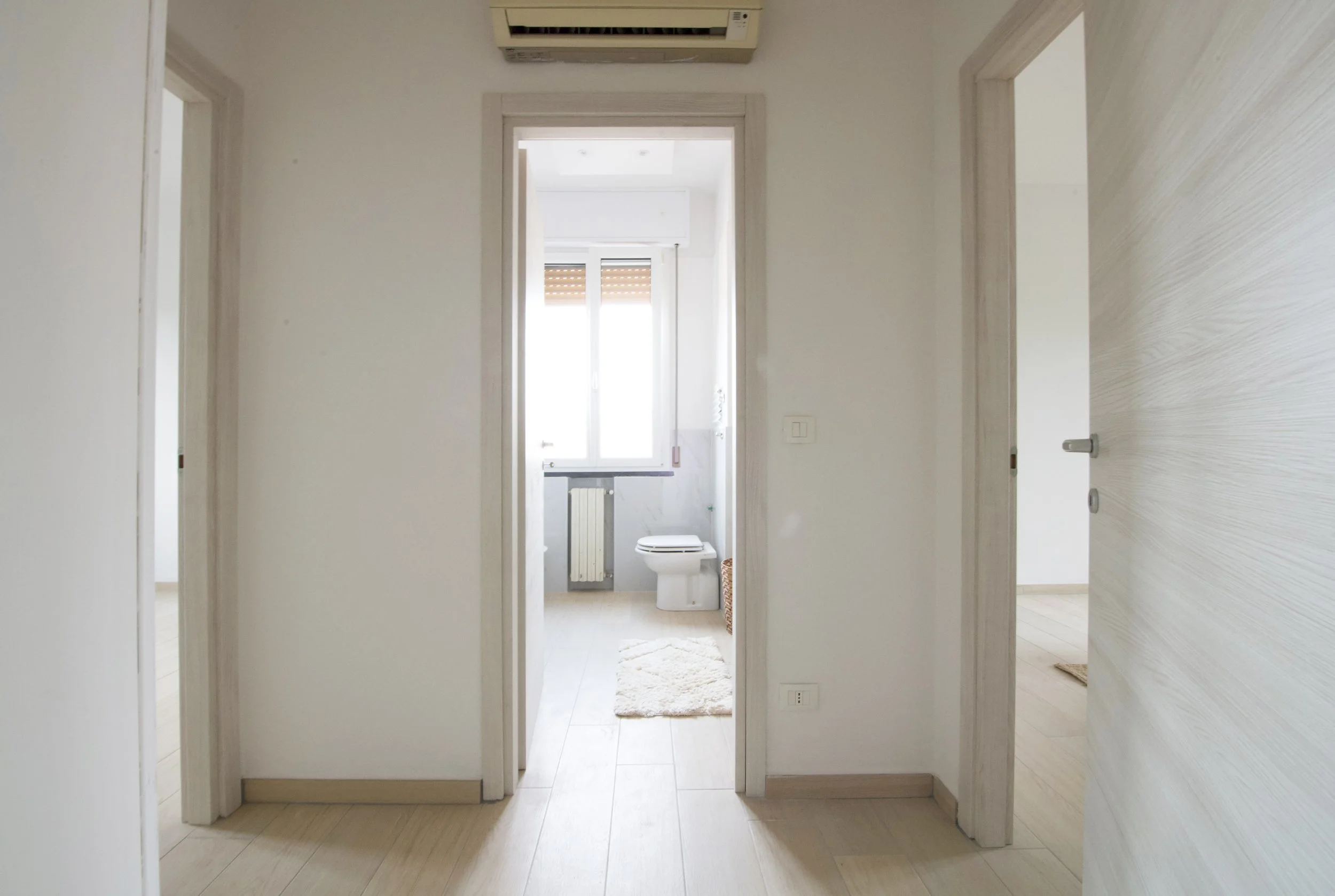 Corridoio di un appartamento con porte aperte, pavimento in legno chiaro e un bagno con finestra e WC visibile al centro.