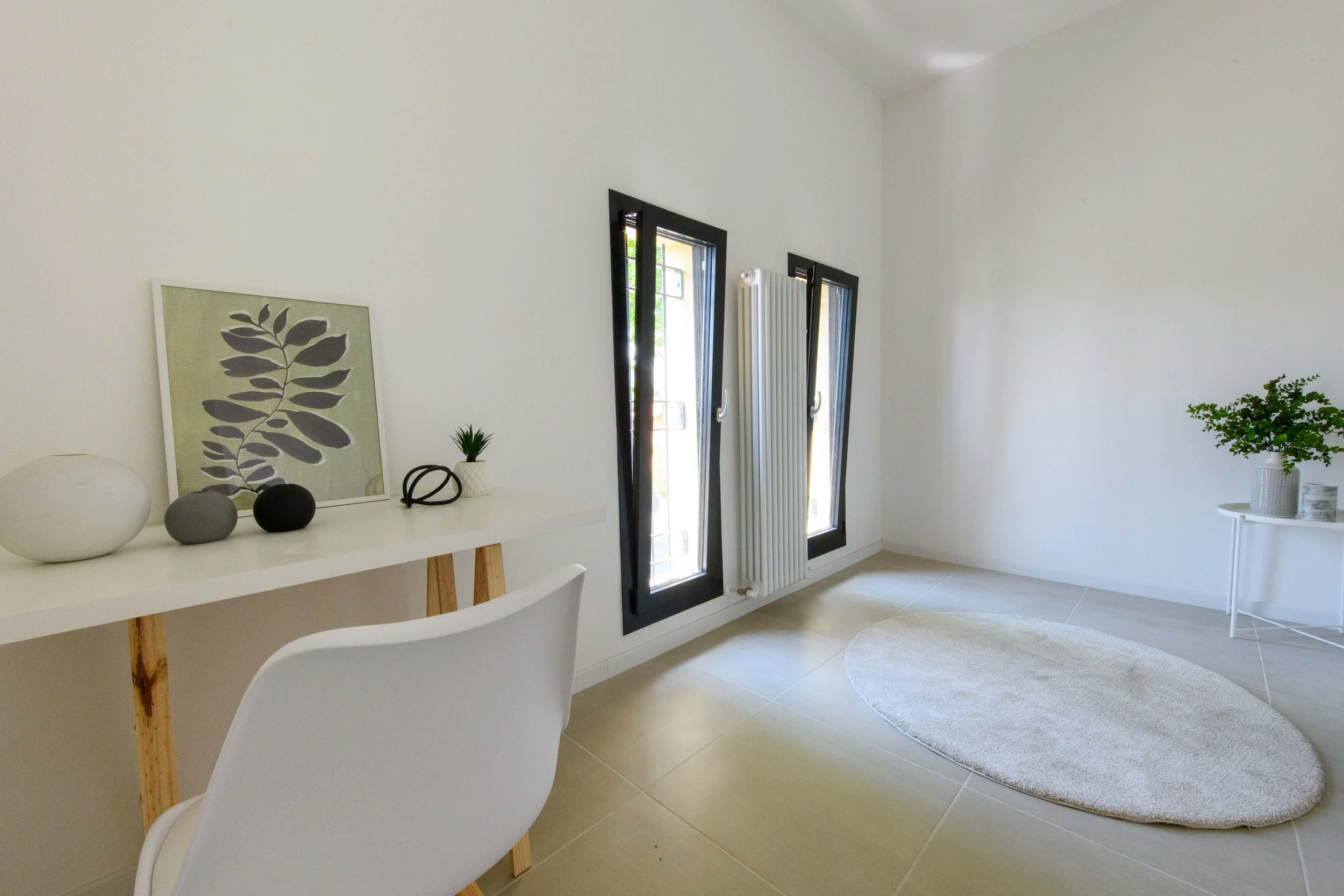 Stanza minimalista con sedia bianca, scrivania con decorazioni, due finestre, un termosifone e tappeto ovale chiaro.