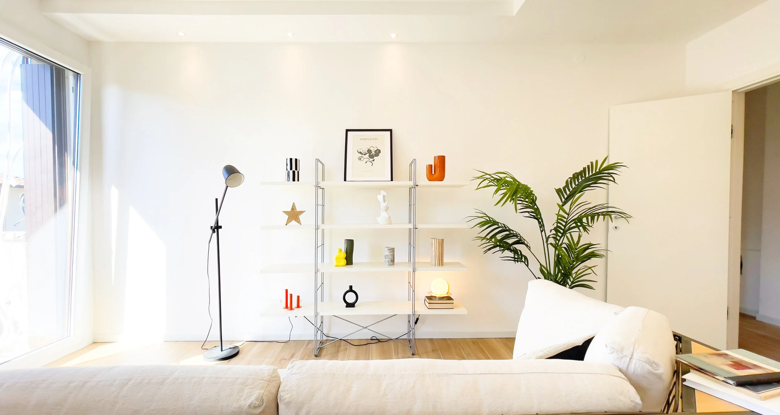 Salotto moderno con scaffale bianco, decorato con oggetti artistici e piante. Lampada da terra nera accanto a divano beige. Luce naturale entra dalle finestre.