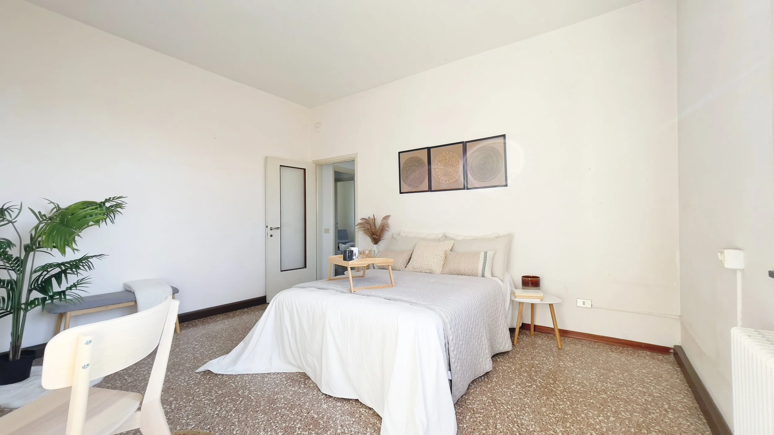 Camera da letto moderna con letto matrimoniale, lenzuola bianche, cuscini decorativi, quadri appesi sopra il letto, pianta in vaso, tavolino di legno e sedia di legno.