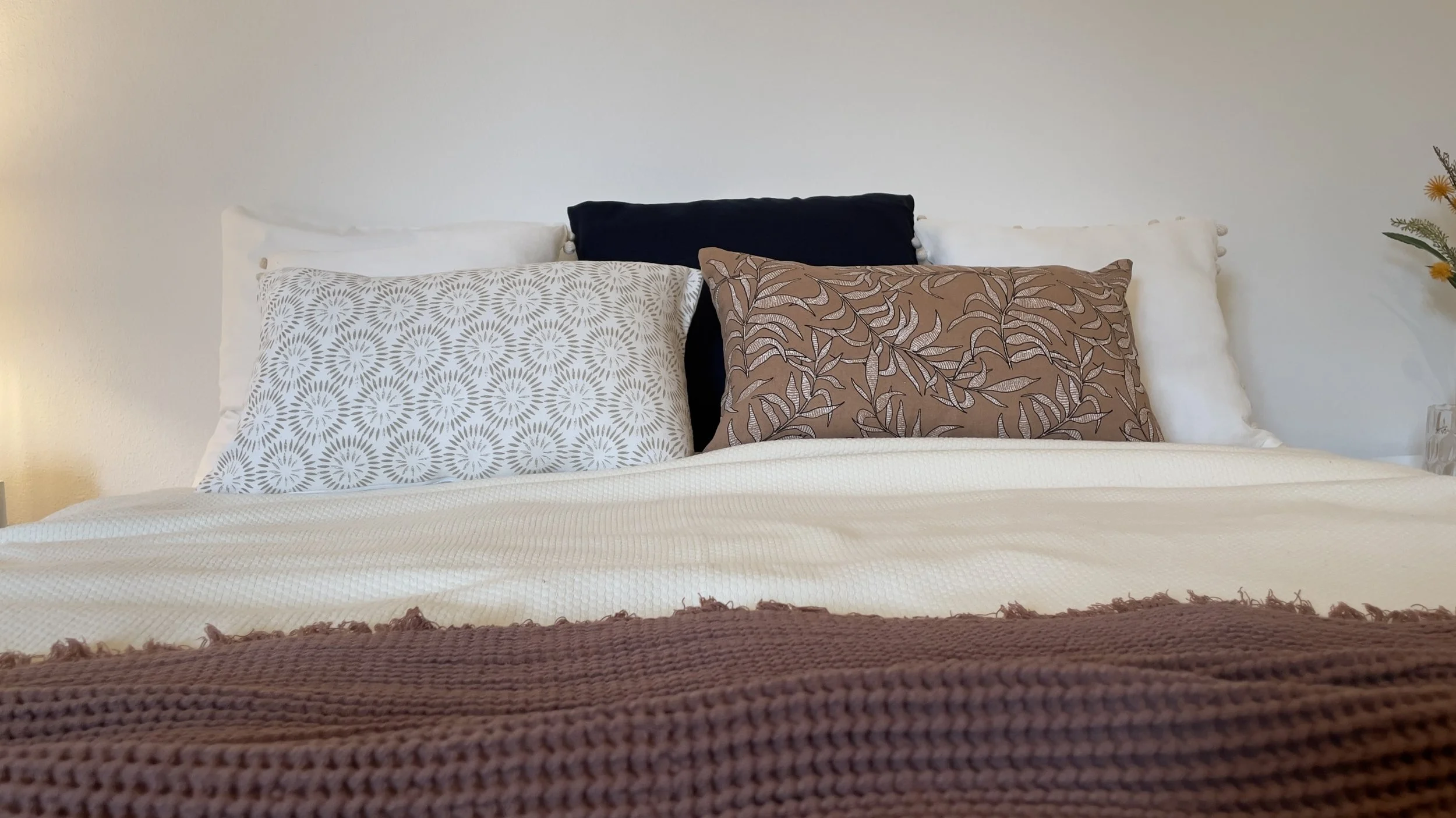Letto con cuscini decorativi, uno bianco con motivi floreali e uno marrone con motivo foglia, coperta marrone in primo piano, sfondo bianco.
