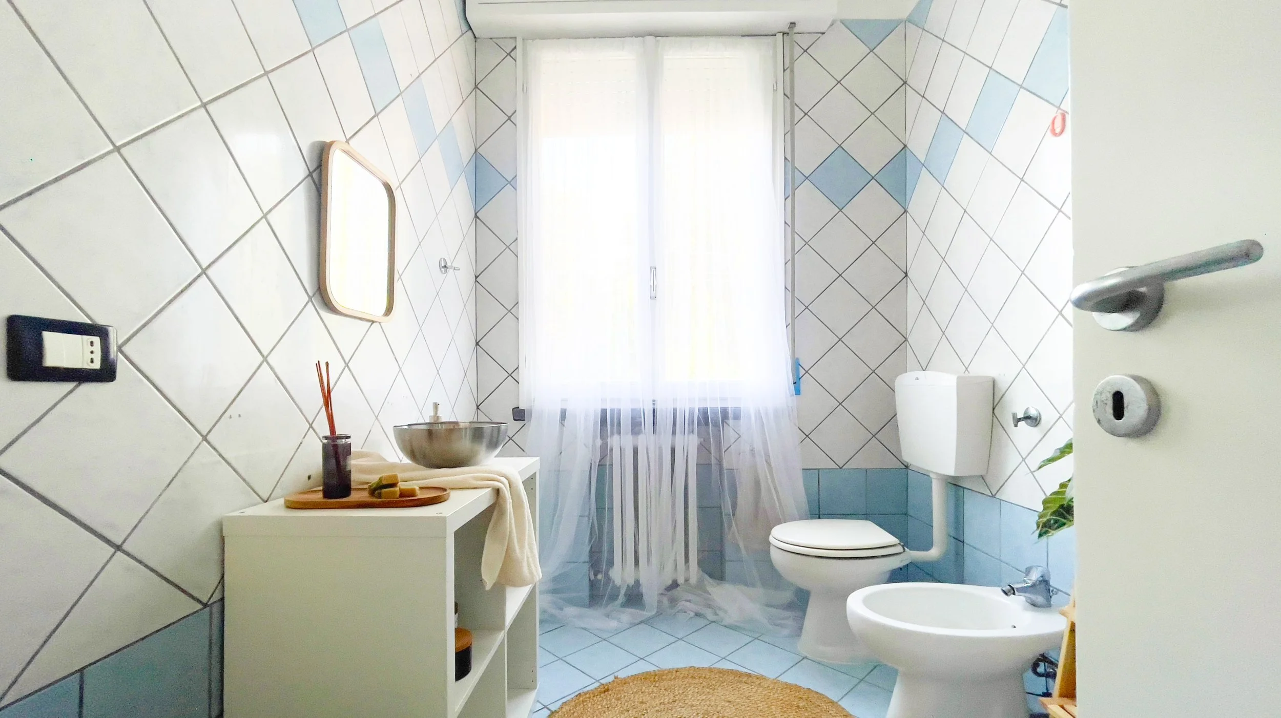 Bagno luminoso con piastrelle bianche e blu, finestra con tenda trasparente, specchio, lavandino, bidet e WC. Mobile bianco con oggetti da bagno e piante decorative.
