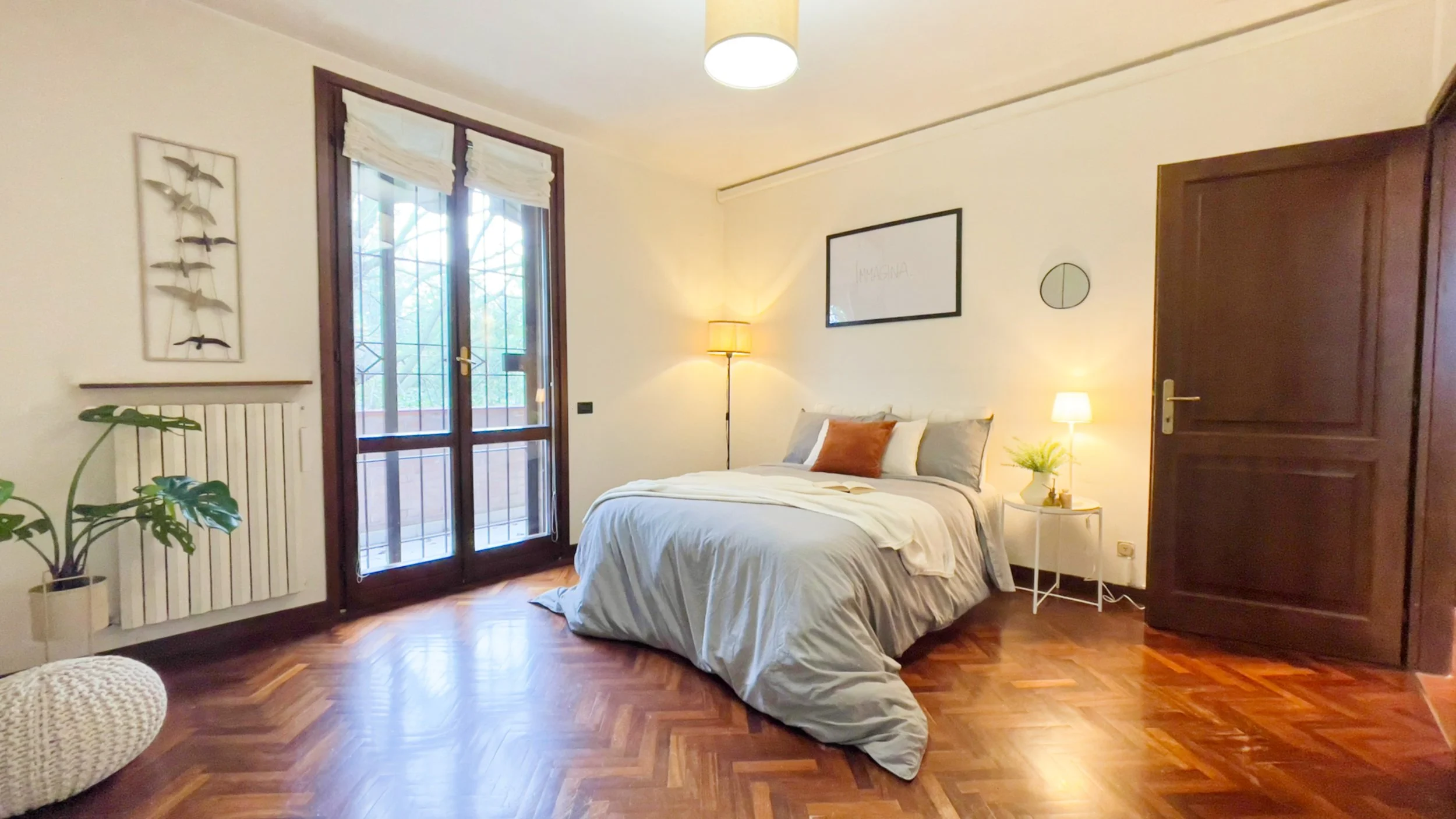 Camera da letto arredata con letto matrimoniale, lampada da terra, comò con pianta e lampada, finestra grande con tende e pavimento in parquet.