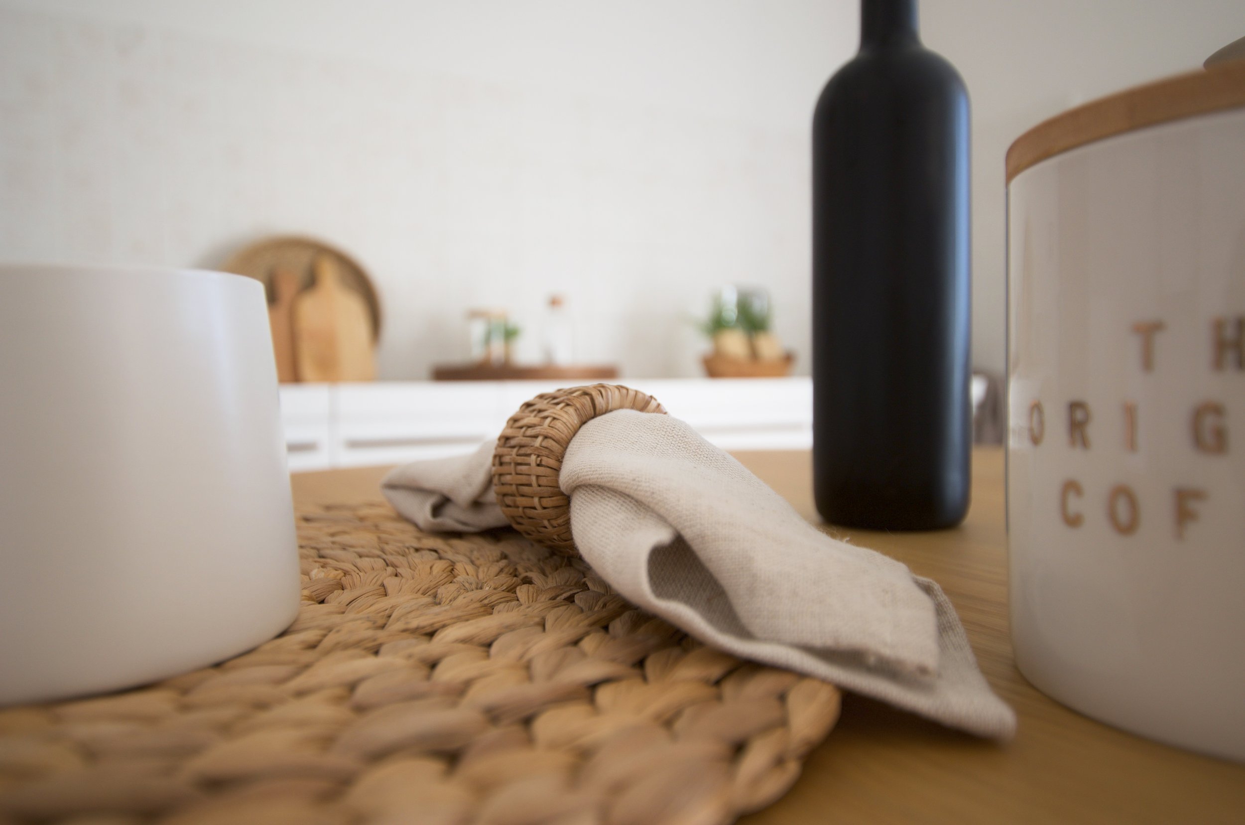 Primo piano di un tavolo da cucina con tovagliolo in tessuto tenuto da un portatovaglioli in rattan su una tovaglietta intrecciata. Sullo sfondo, una bottiglia scura, un barattolo con scritta "The Original Coffee" e taglieri di legno appoggiati contr