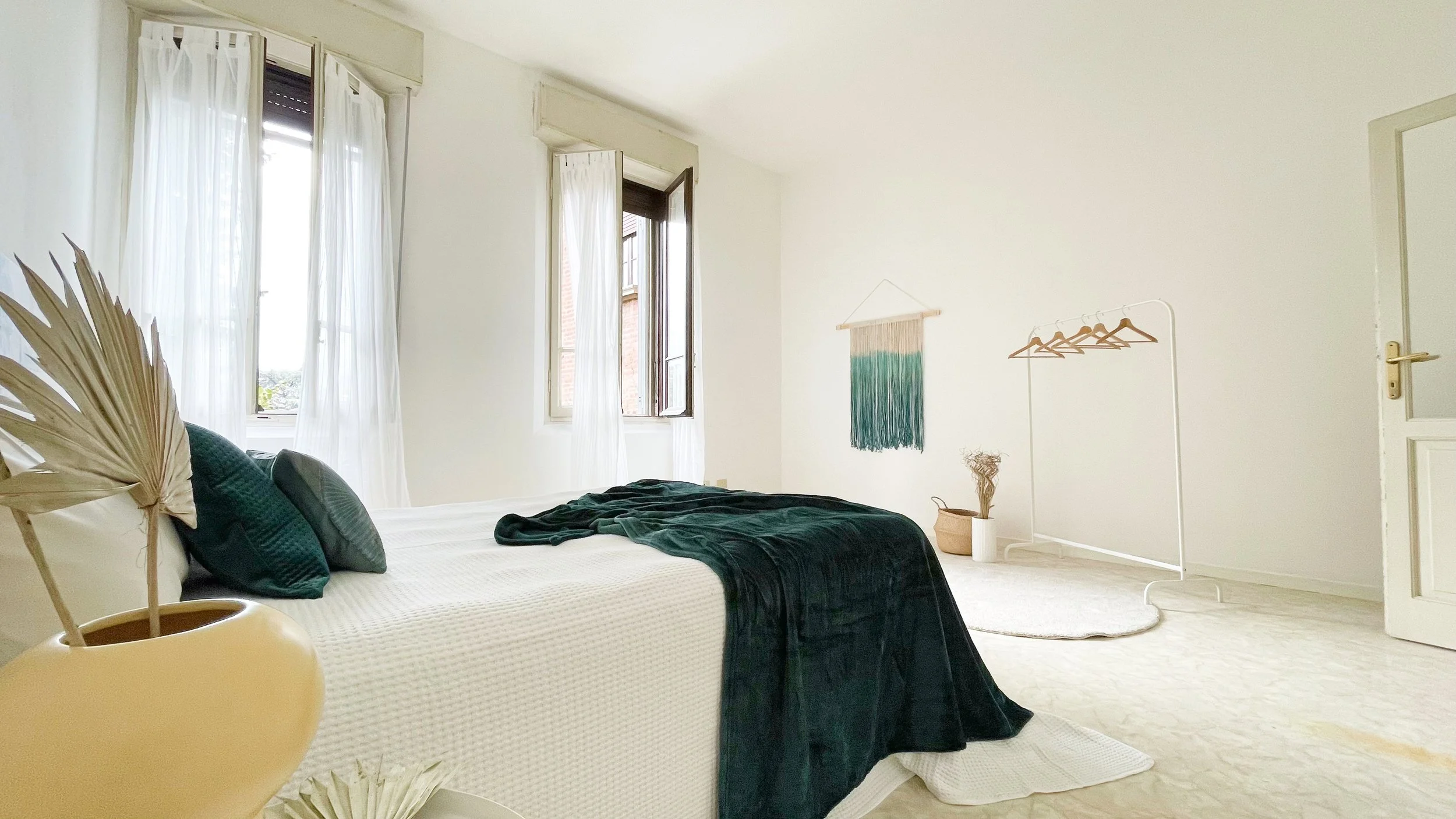 Camera da letto luminosa con letto matrimoniale, coperte verde scuro, finestre con tende bianche, appendiabiti bianco e decorazioni a parete.