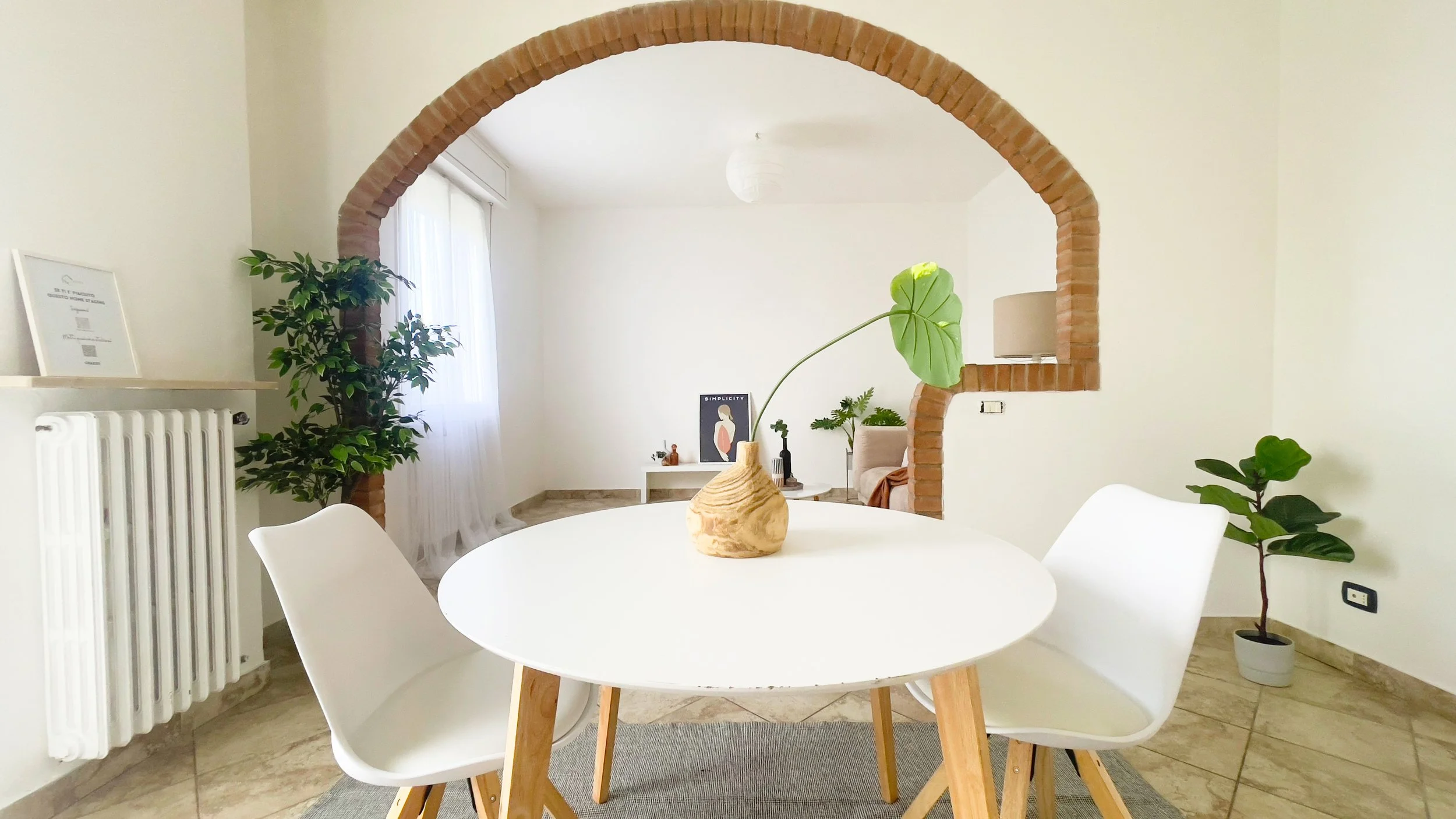 Stanza moderna con tavolo rotondo bianco, sedie bianche, vaso con foglia, arco in mattoni, piante, e arredamento minimalista.