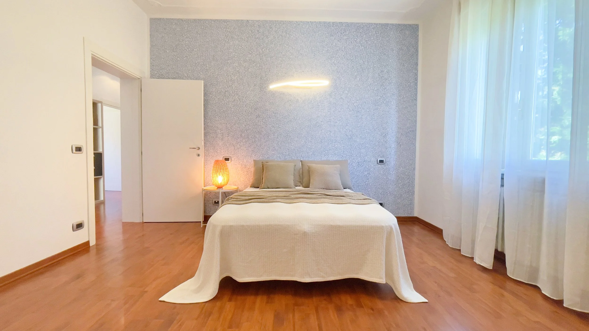 Camera da letto luminosa con letto matrimoniale, copriletto bianco, e lampada sul comodino. Pavimento in legno e parete con lampada moderna sopra il letto.