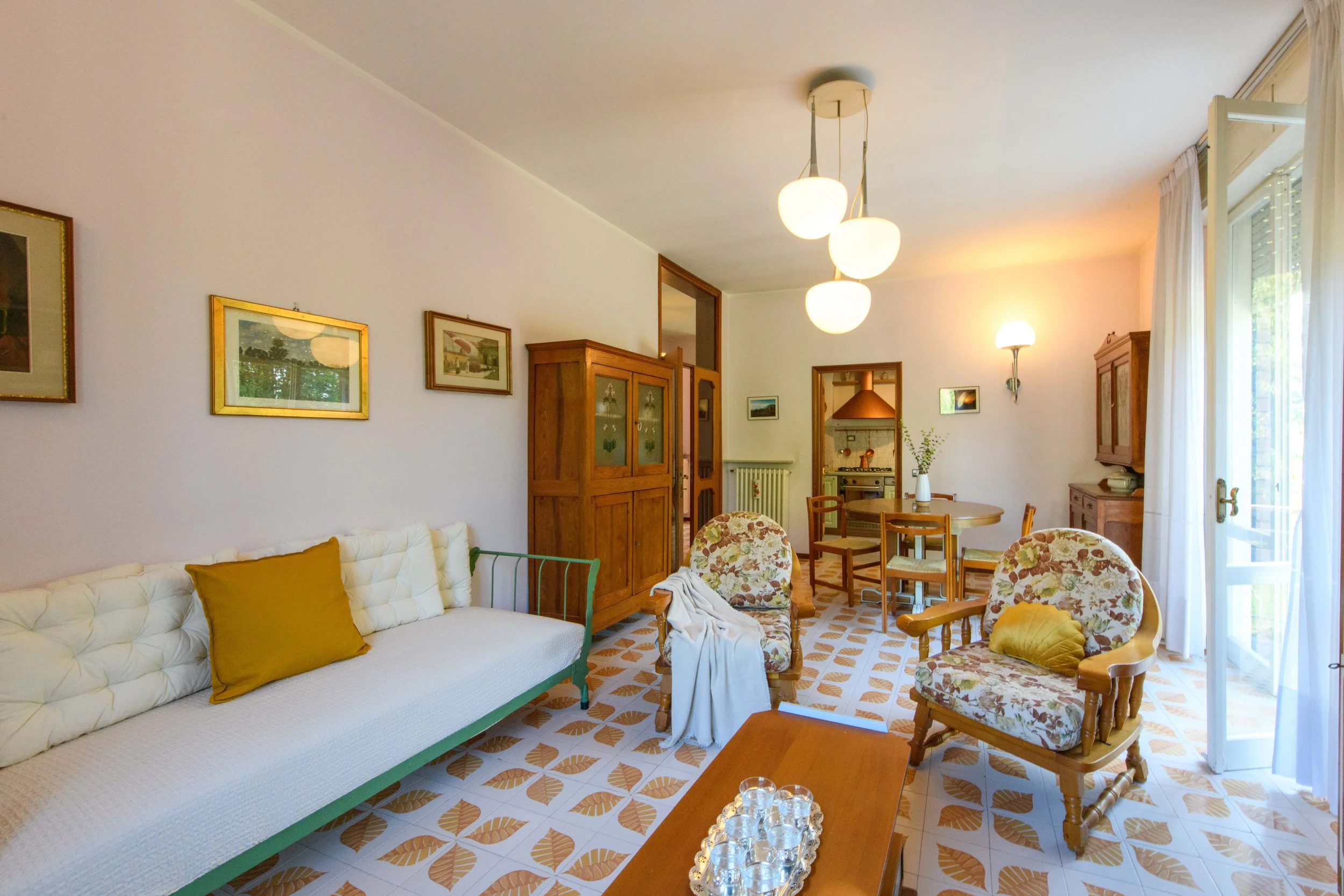 Soggiorno luminoso con arredamento vintage, divano bianco con cuscino giallo, due sedie con stoffa floreale, tavolo in legno, lampade a soffitto e quadri appesi alle pareti. Cucina visibile sullo sfondo tramite una porta aperta.