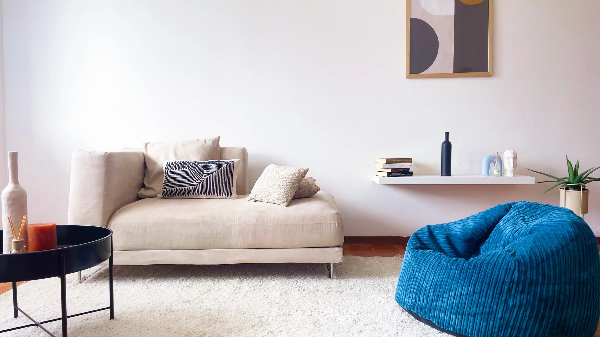 Soggiorno moderno con divano beige, cuscini decorativi, tavolo rotondo nero con candele, parete bianca con mensola galleggiante, libri, vaso nero, bean bag blu e pianta in vaso, quadro astratto appeso.