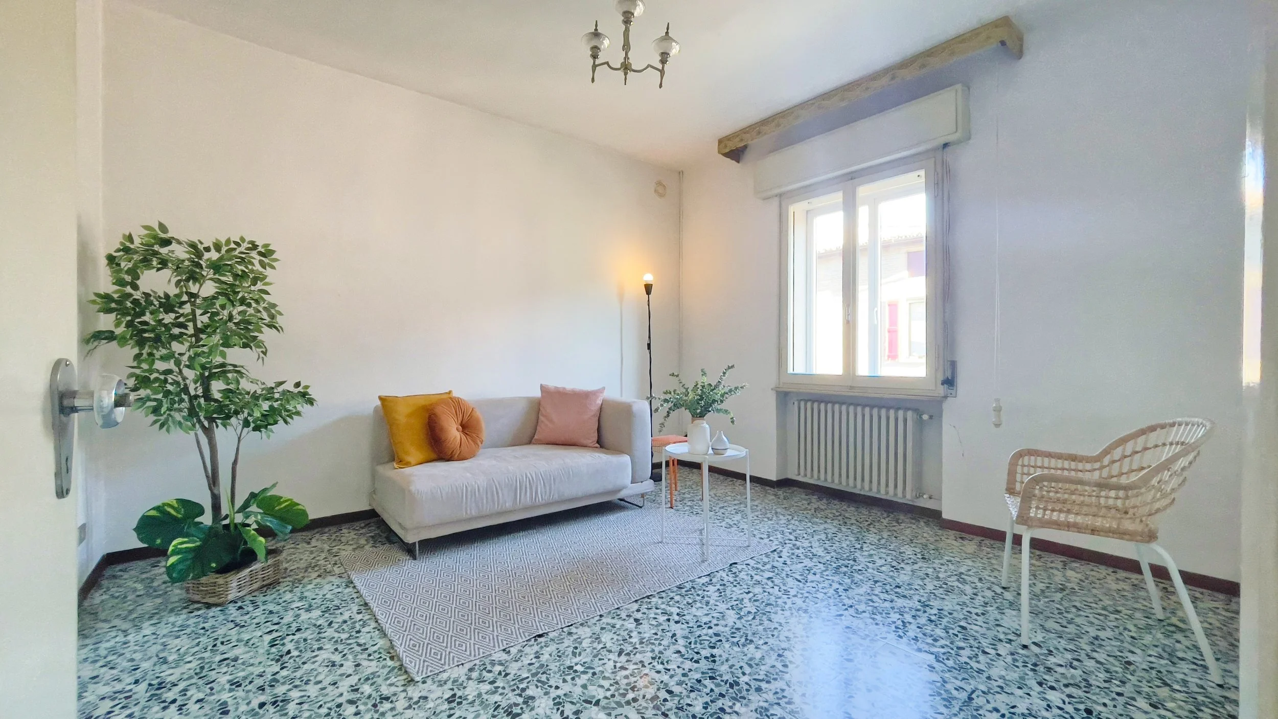 Soggiorno con divano beige, cuscini colorati, tavolino rotondo bianco, pianta decorativa in vaso, sedia intrecciata, pavimento in graniglia e grande finestra.