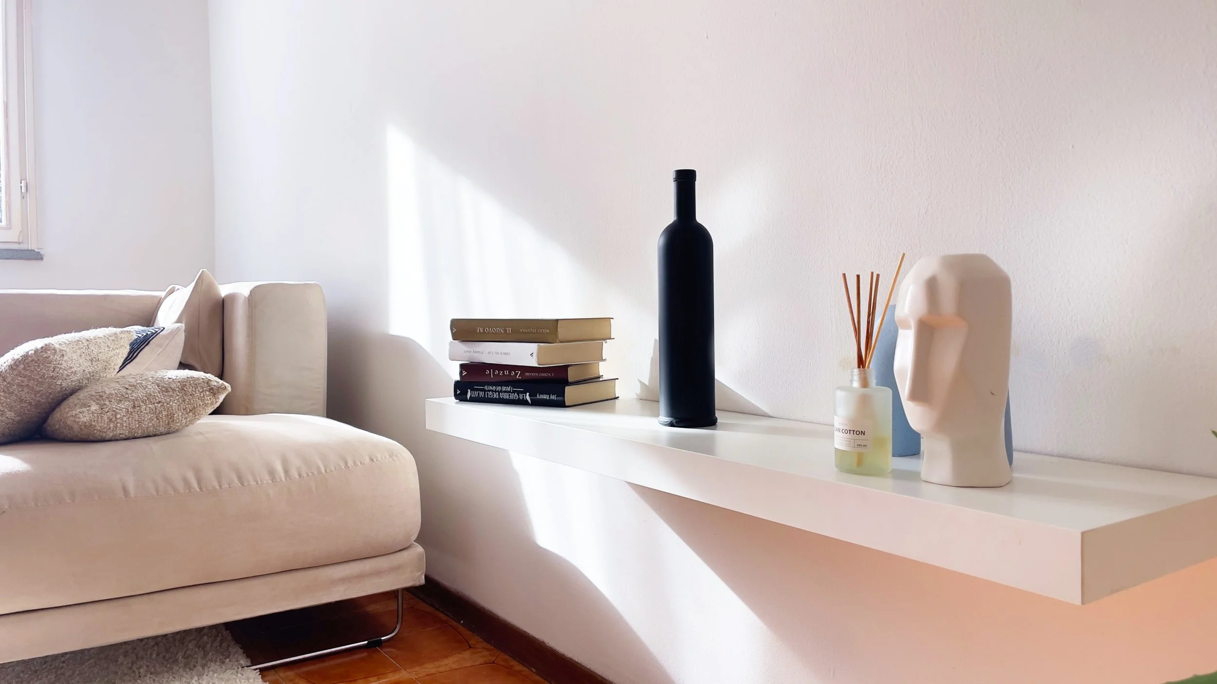 Interno di una stanza con divano beige, cuscini decorativi, mensola bianca con libri, una bottiglia nera, un diffusore di fragranze e un vaso decorativo a forma di testa.