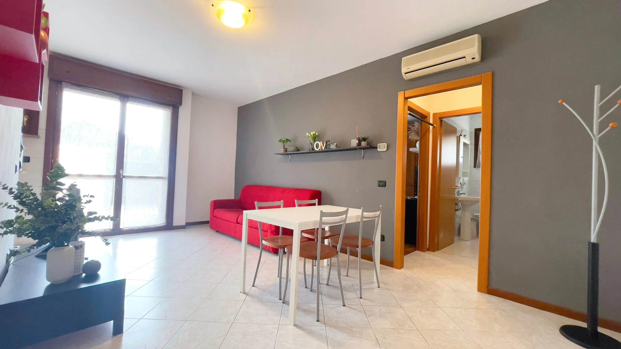 Una sala moderna con pareti grigie, un divano rosso, tavolo e sedie bianche, mensola con oggetti decorativi. Sul lato sinistro, una grande finestra e sul destro, una porta aperta verso il bagno. Un appendiabiti è visibile nell'angolo destro.