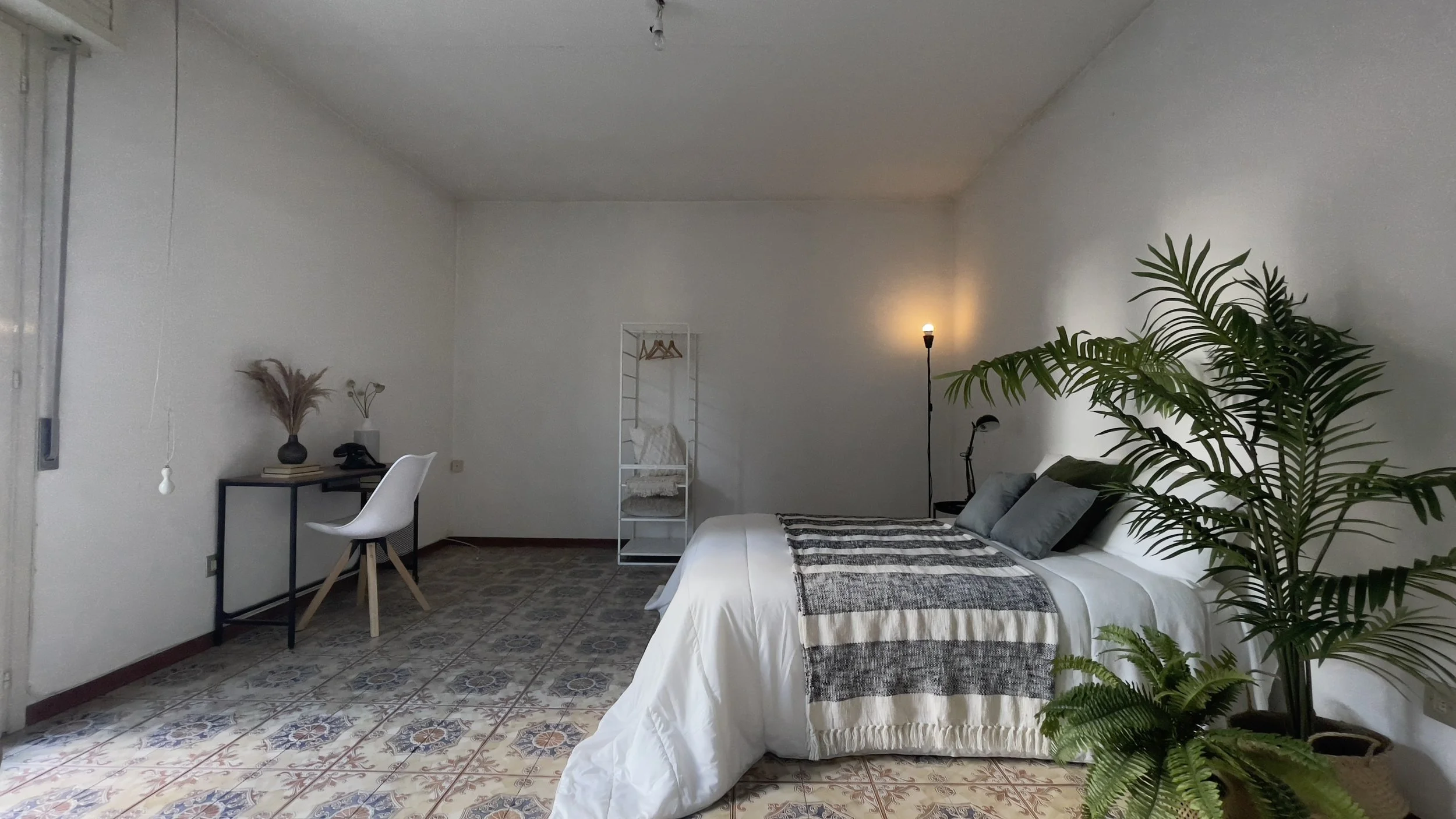 Camera da letto minimalista con letto matrimoniale, piante verdi, lampada da terra, scrivania con sedia bianca, e stand appendiabiti. Pavimento con piastrelle decorative.
