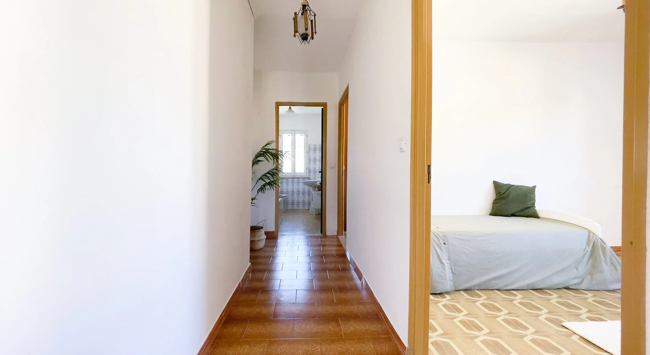 Interno di una casa con un corridoio, una camera da letto a destra con un letto e un cuscino verde, e un bagno alla fine del corridoio. Un vaso con una pianta si trova nel corridoio. Le pareti sono bianche e il pavimento è di piastrelle.