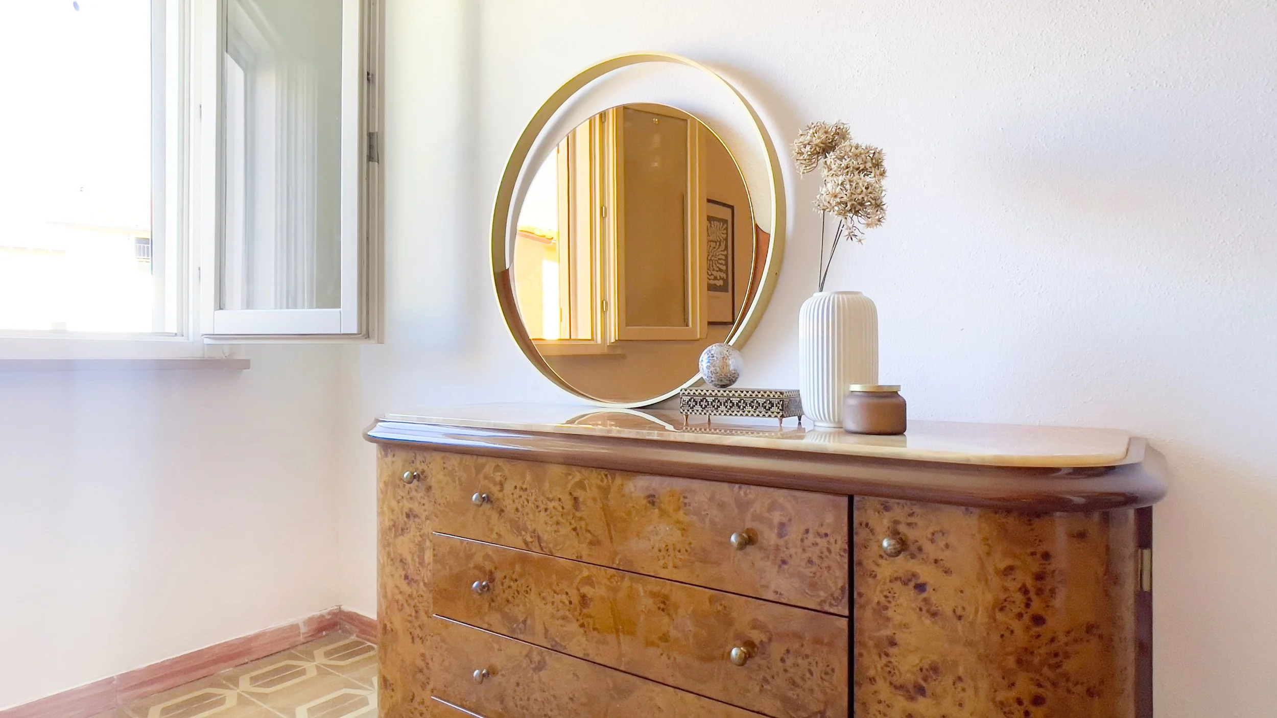 Comò in legno con specchio rotondo, vaso bianco con fiori secchi e una scatola decorativa. A fianco, una finestra aperta.