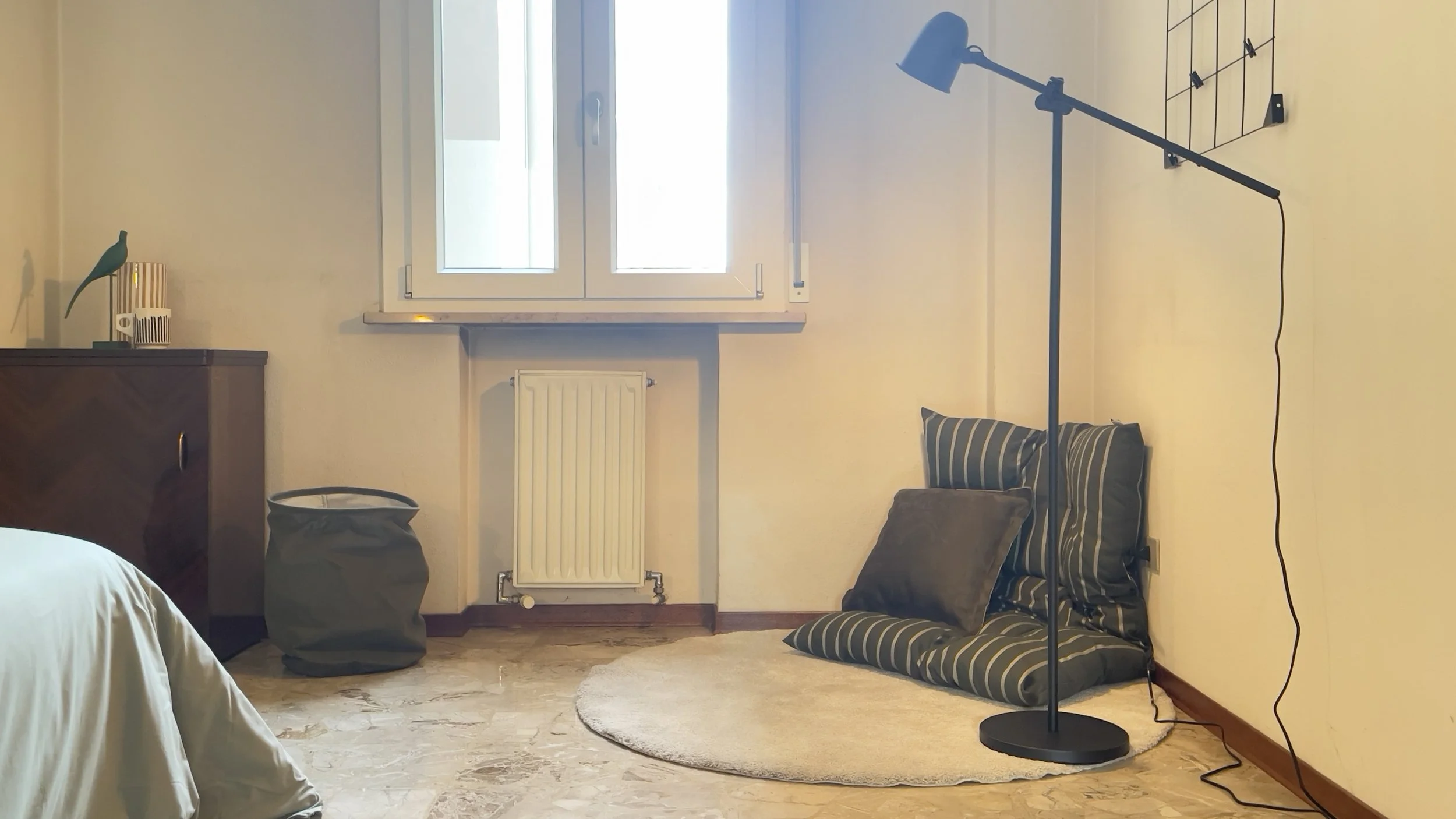 Soggiorno moderno con cuscini a righe sul pavimento, lampada da terra nera, finestra con cornice bianca, mobile basso con decorazioni e un cesto verde.