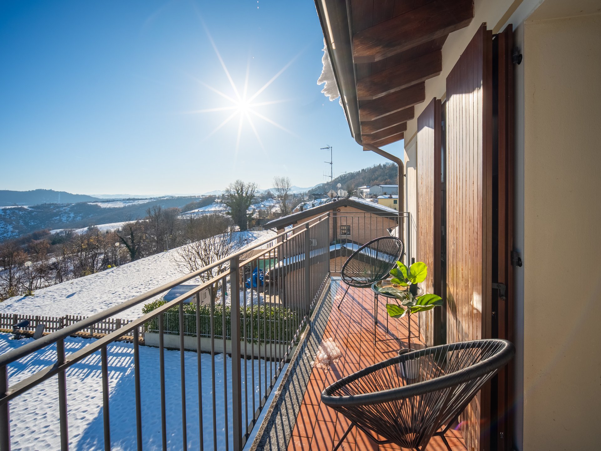 Balcone soleggiato con due sedie e piante, paesaggio innevato e cielo sereno sullo sfondo.