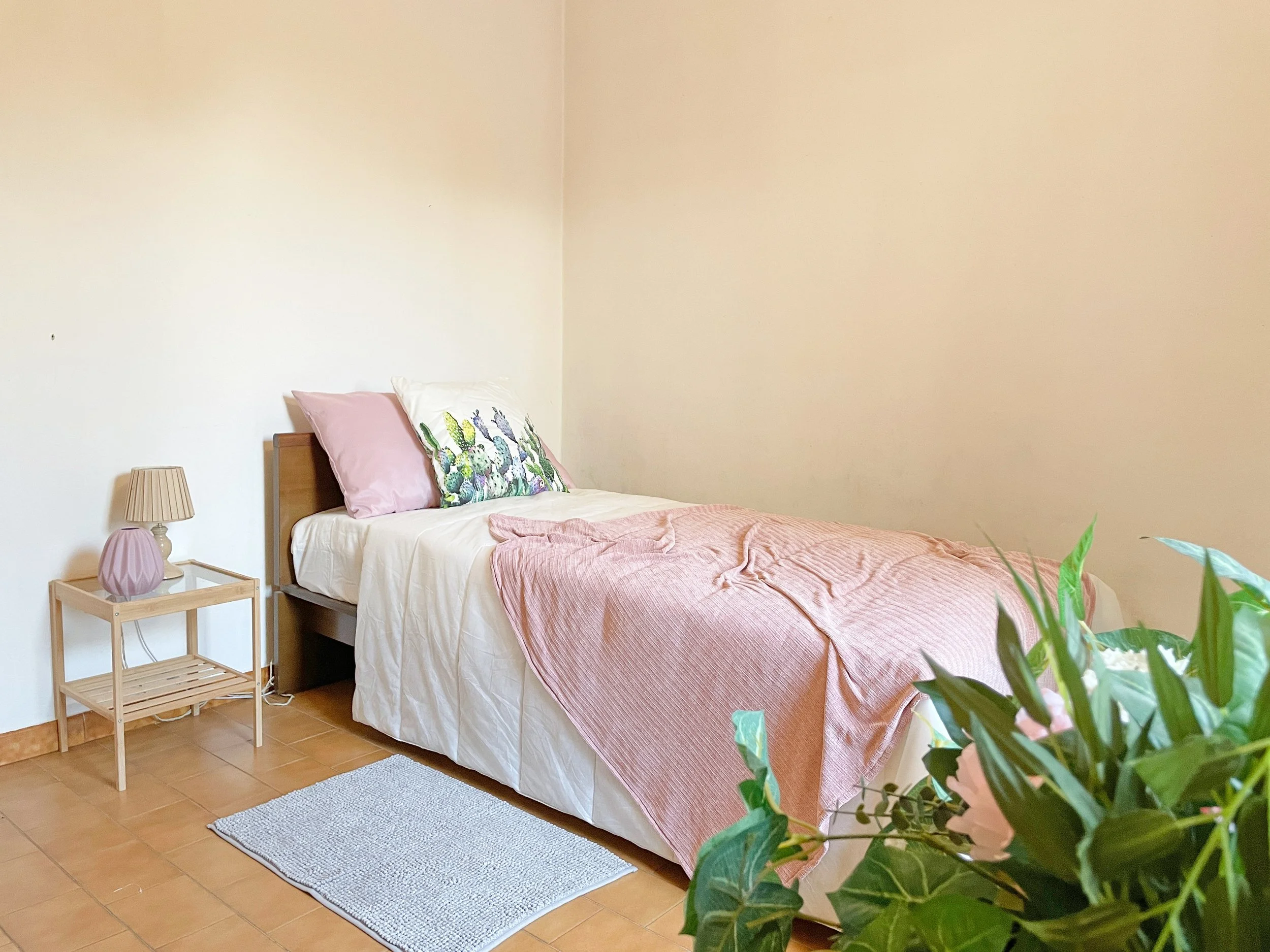 Camera da letto con letto singolo, biancheria rosa, lampada su tavolino e pianta decorativa.
