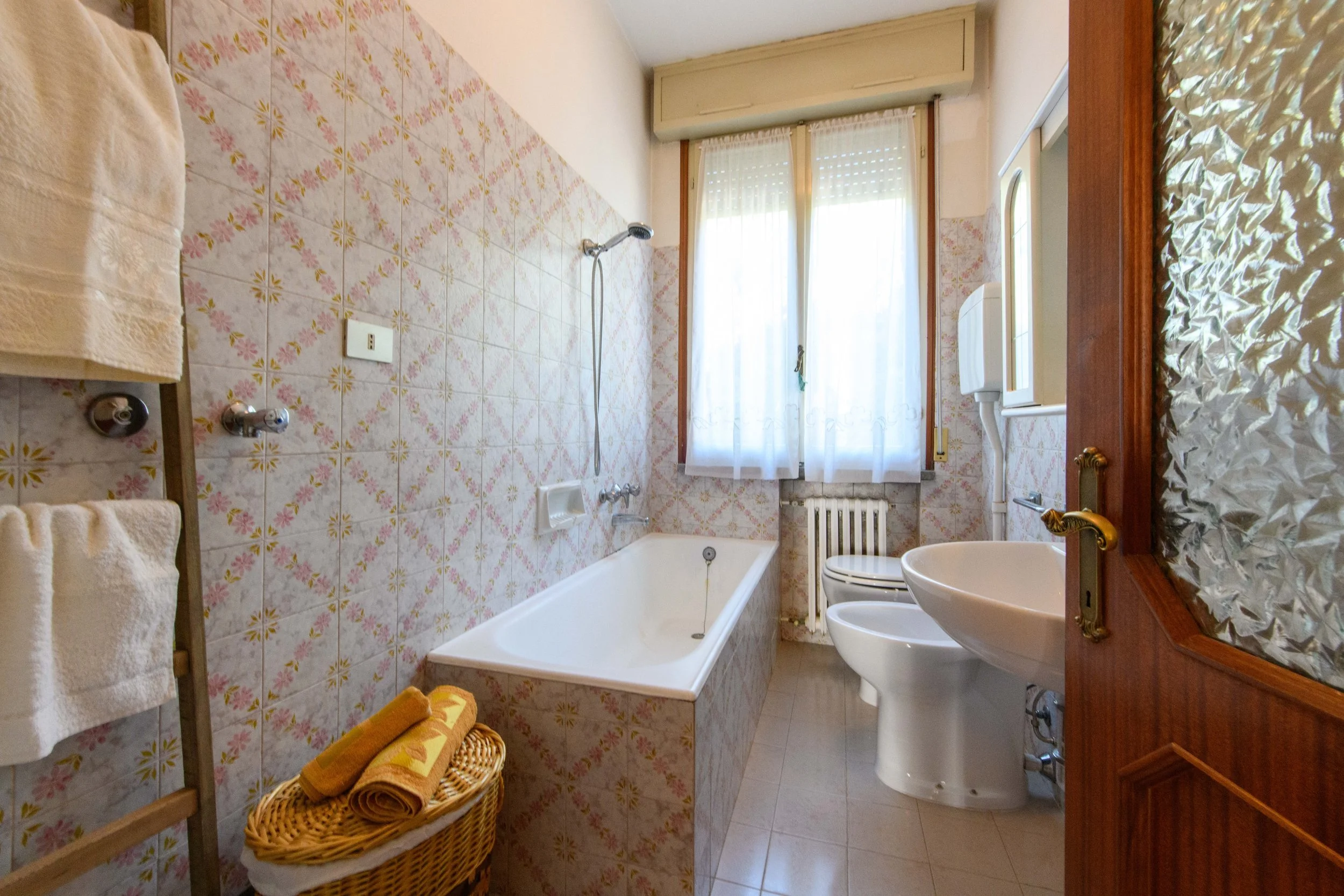 Bagno con vasca bianca, lavandino, bidet e water; rivestimenti con piastrelle a motivi floreali e asciugamani appesi. Cesto con asciugamani vicino alla vasca. Finestra con tende bianche e porta in legno con vetro decorato.