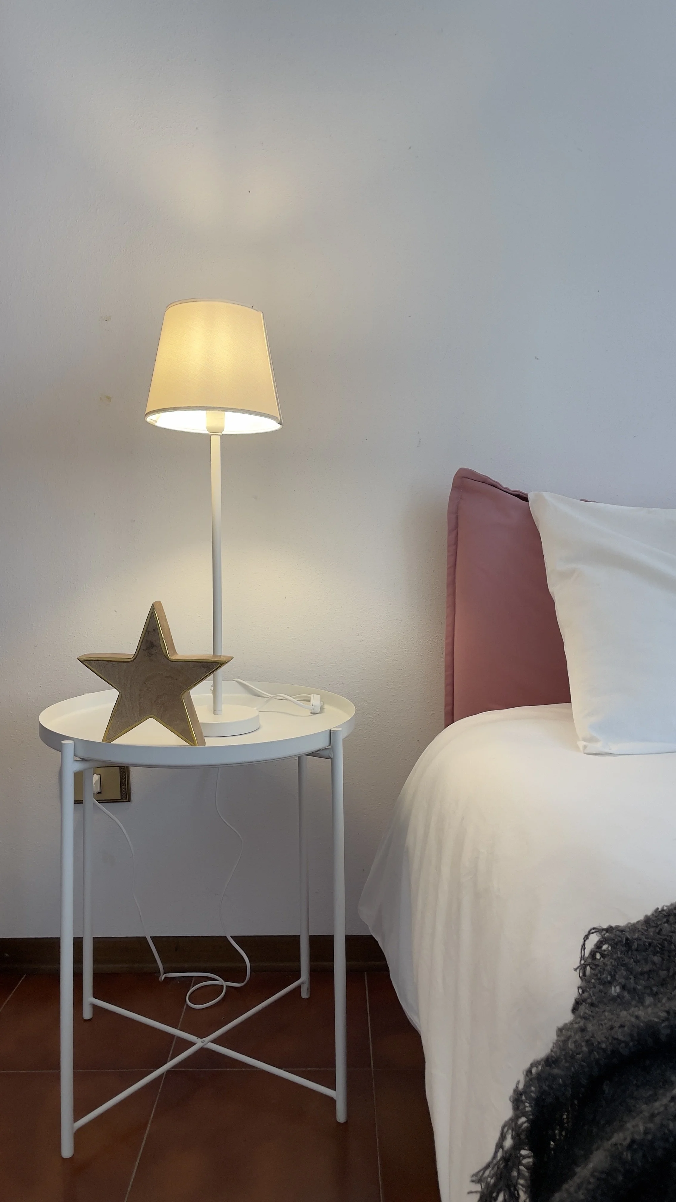Camera da letto con un comodino bianco, una lampada accesa e una decorazione a forma di stella. Accanto c'è un letto con cuscini bianchi e una testiera rosa.