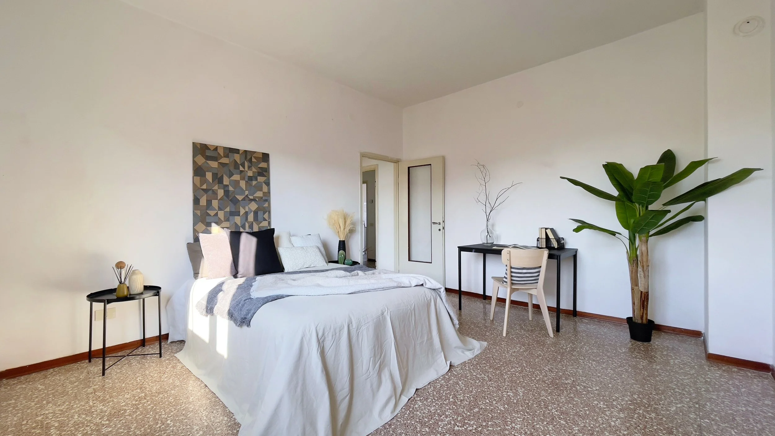 Camera da letto moderna con letto matrimoniale, lenzuola bianche e cuscini, tavolino nero con decorazioni, pianta alta, scrivania nera con sedia chiara, parete bianca e pavimento in piastrelle.