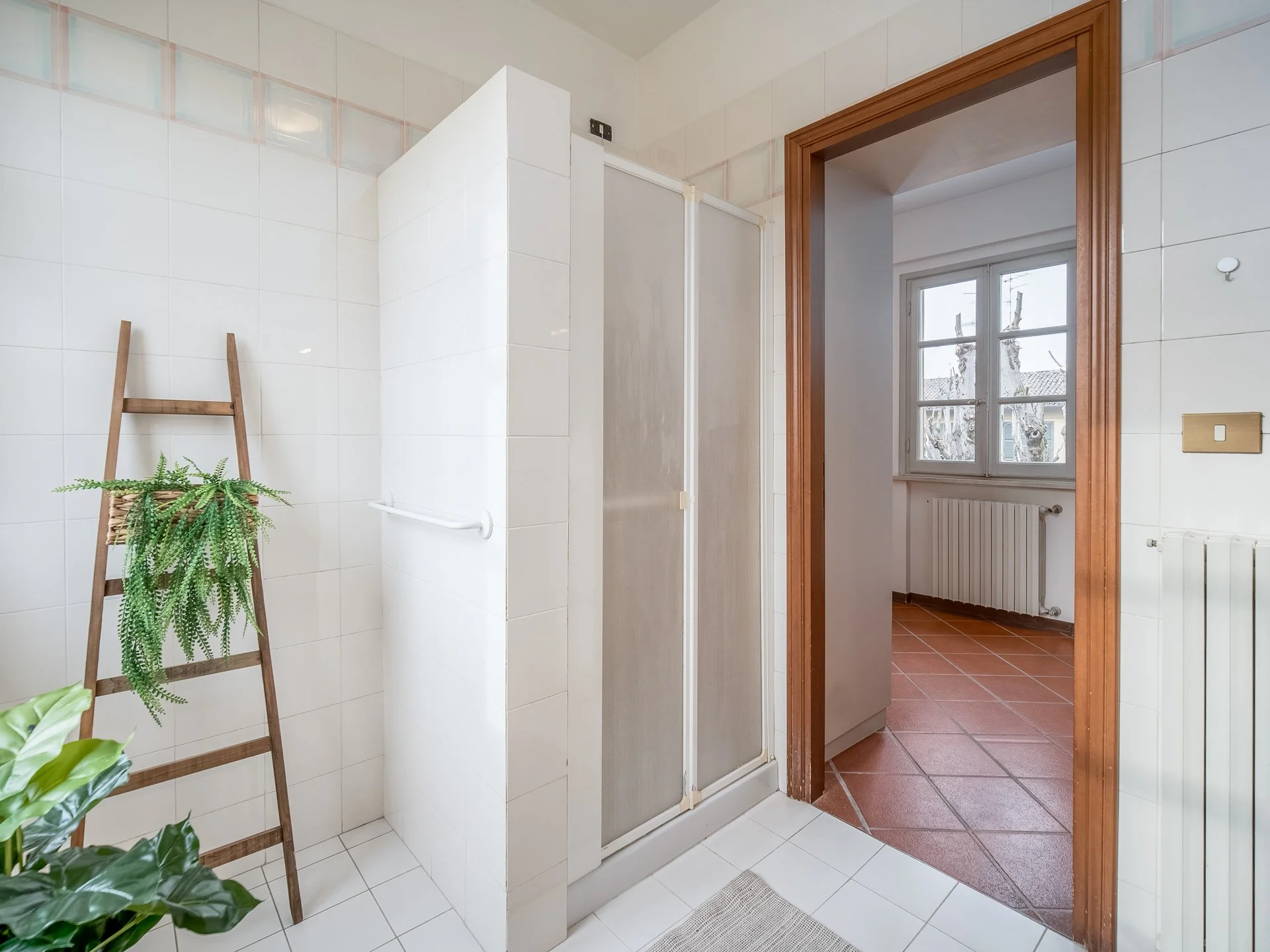 Bagno moderno con doccia, piastrelle bianche, scala in legno con pianta verde e porta verso una stanza con pavimento in cotto.