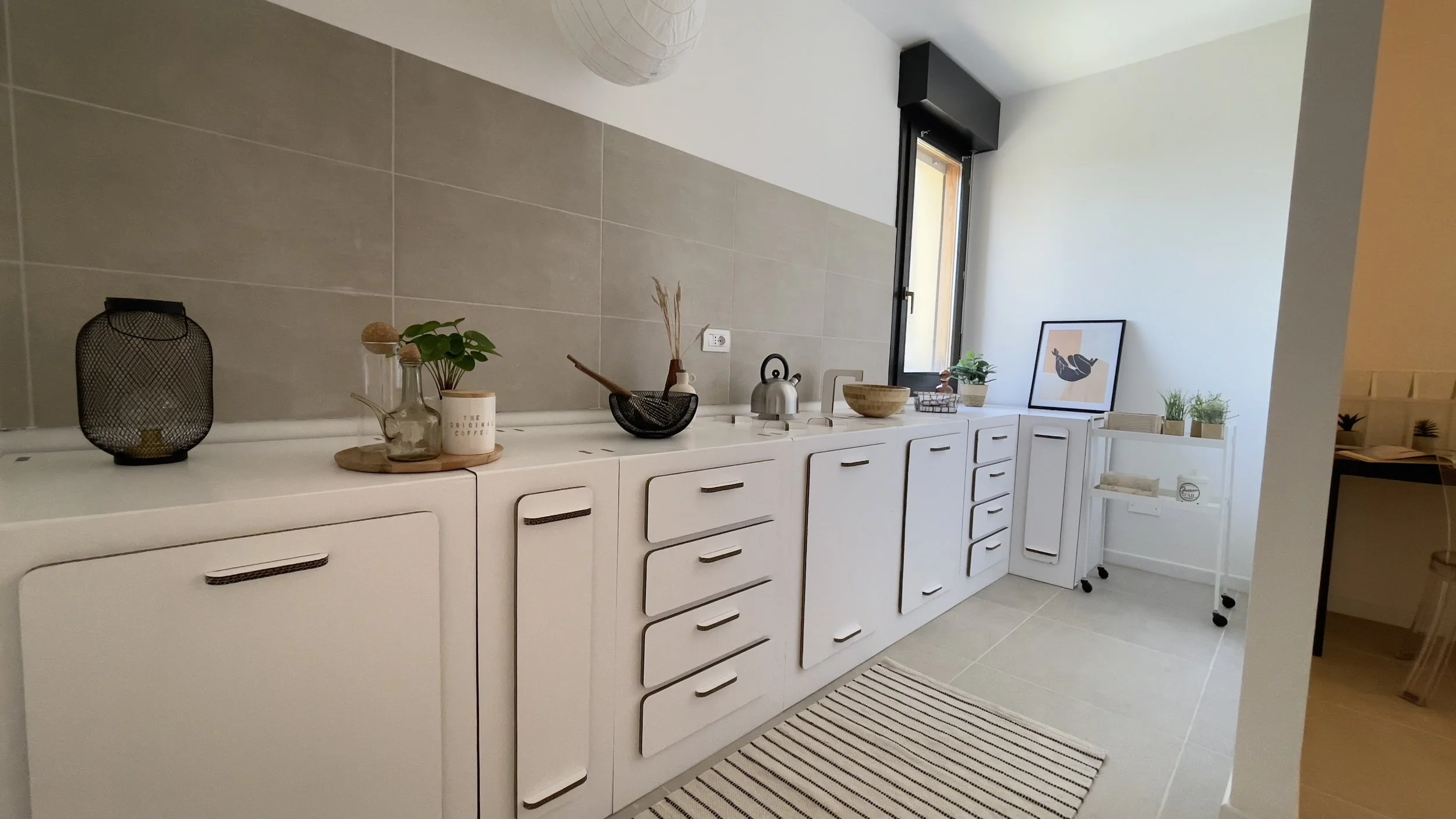 Cucina minimalista con mobili bianchi, decorazioni semplici, pianta, bollitore, finestra e tappeto a righe.