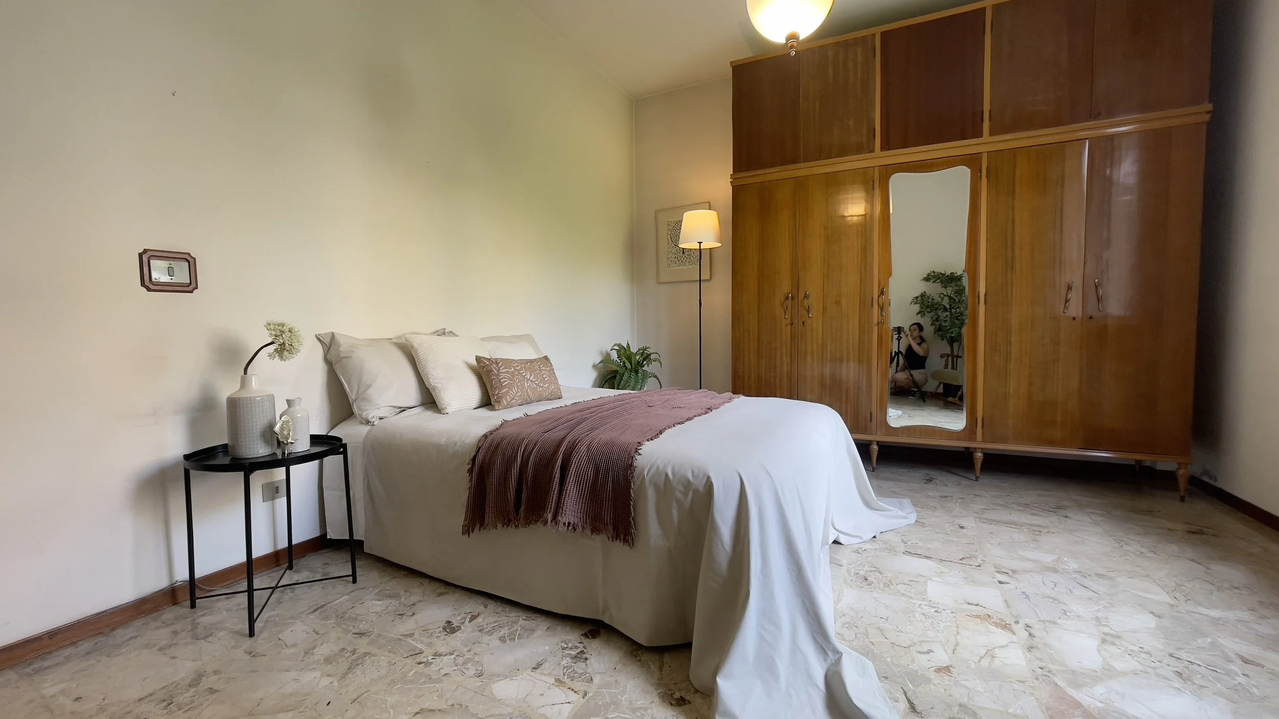 Camera da letto con letto matrimoniale, comodino con vasi, armadio specchiato, pavimento in marmo, lampada a stelo e pianta decorativa.