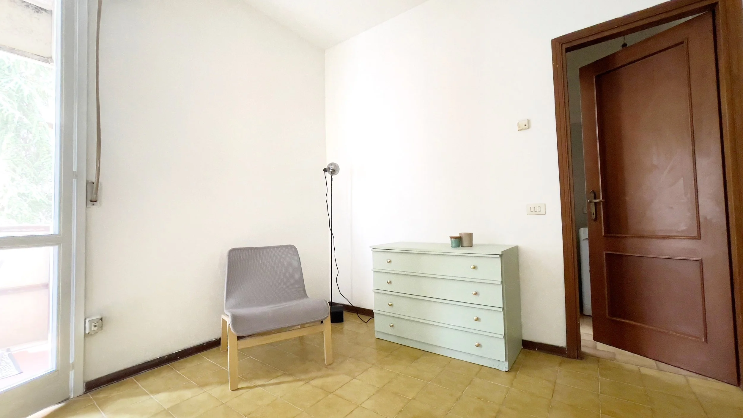 Stanza con una sedia grigia, una lampada da terra nera, un cassettone verde chiaro e una porta di legno aperta.