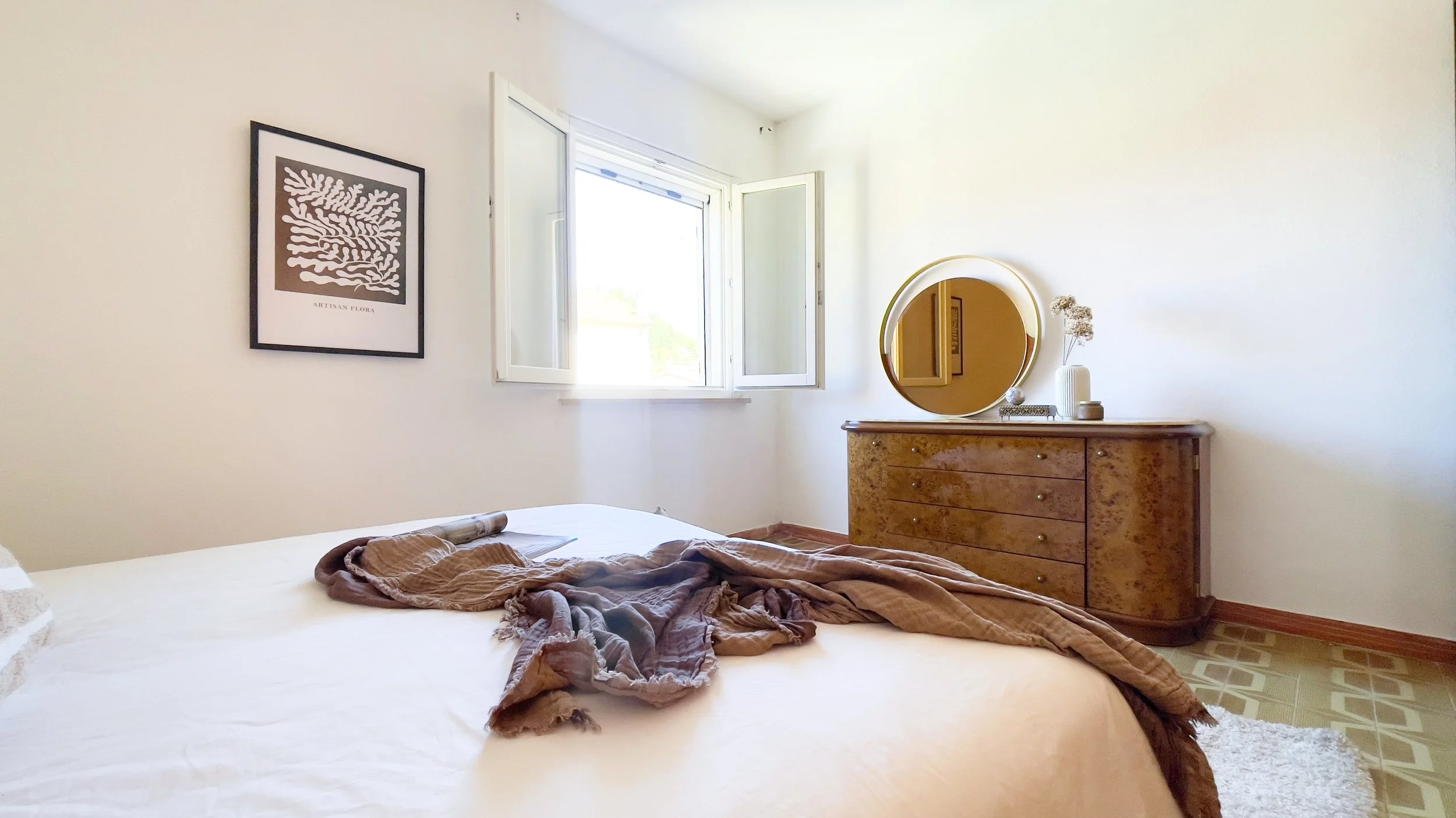 Camera da letto luminosa con letto matrimoniale, coperta marrone gettata sopra, finestra aperta sulla parete sinistra, quadro decorativo, e comò in legno con specchio rotondo e vasi decorativi.