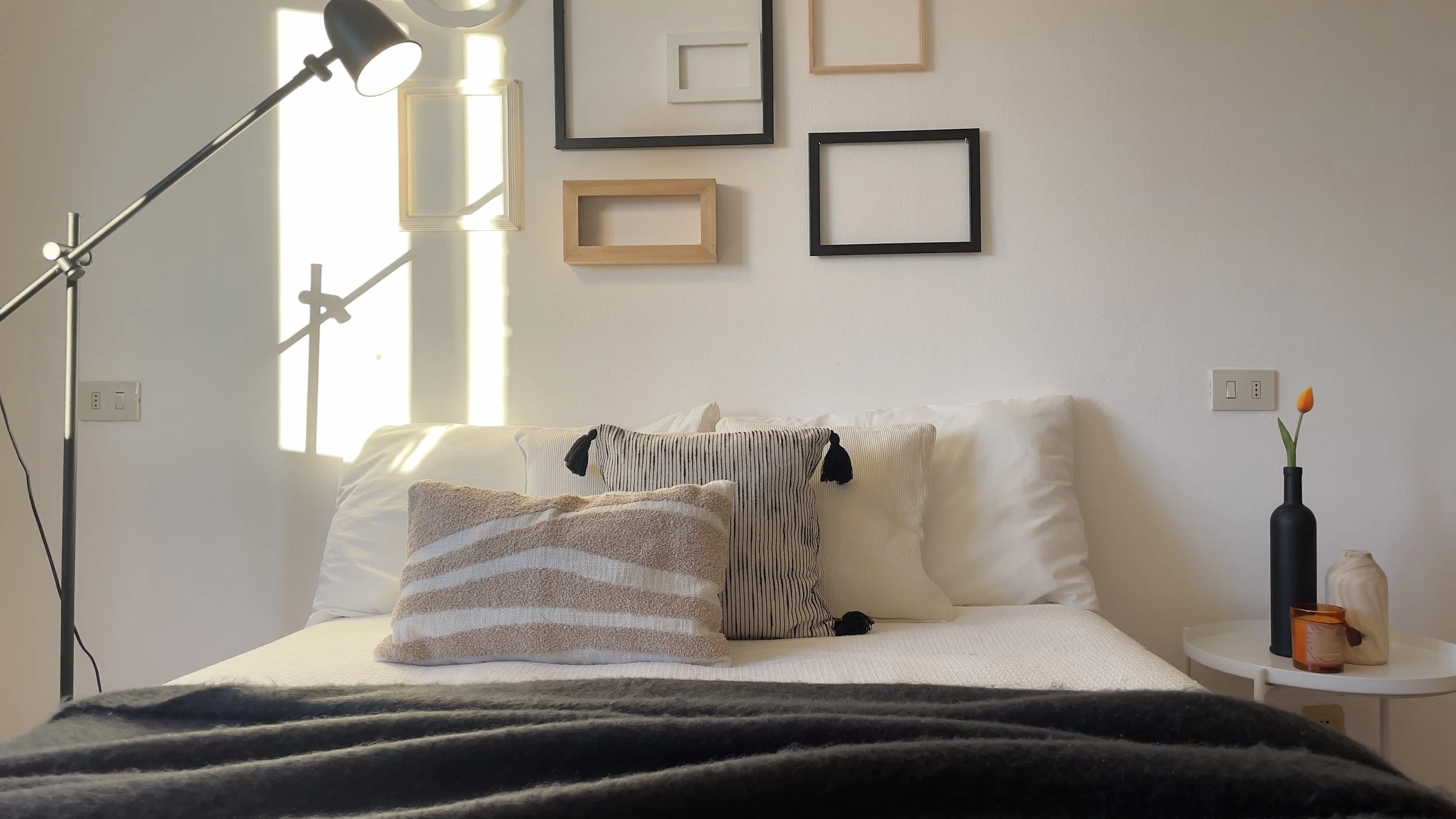 Camera da letto con letto, cuscini, lampada, cornici decorative e tavolino con vaso e pianta.