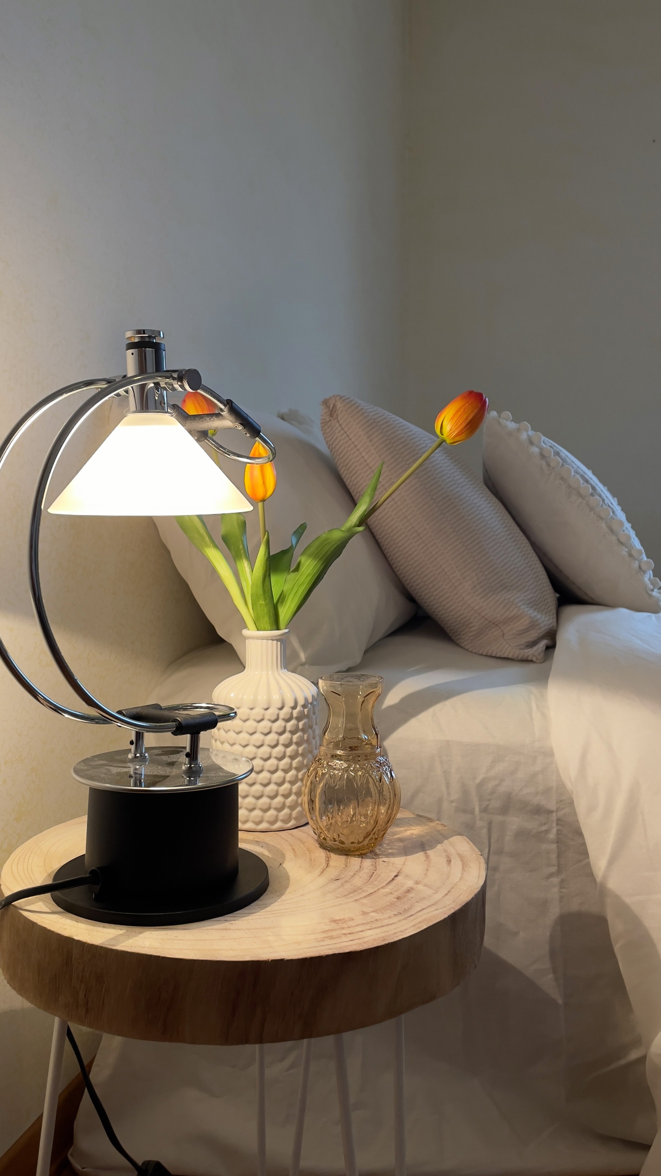 Comodino con lampada moderna, vaso bianco con tulipani arancioni e brocca di vetro, vicino a un letto con cuscini e lenzuola bianche.