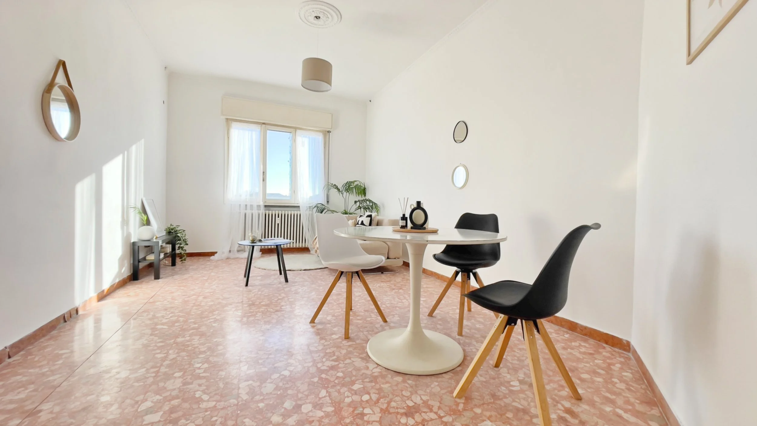 Stanza luminosa con tavolo rotondo bianco, quattro sedie moderne, pareti bianche con specchi decorativi, pavimento in marmo rosa, piante e una grande finestra con tende trasparenti.