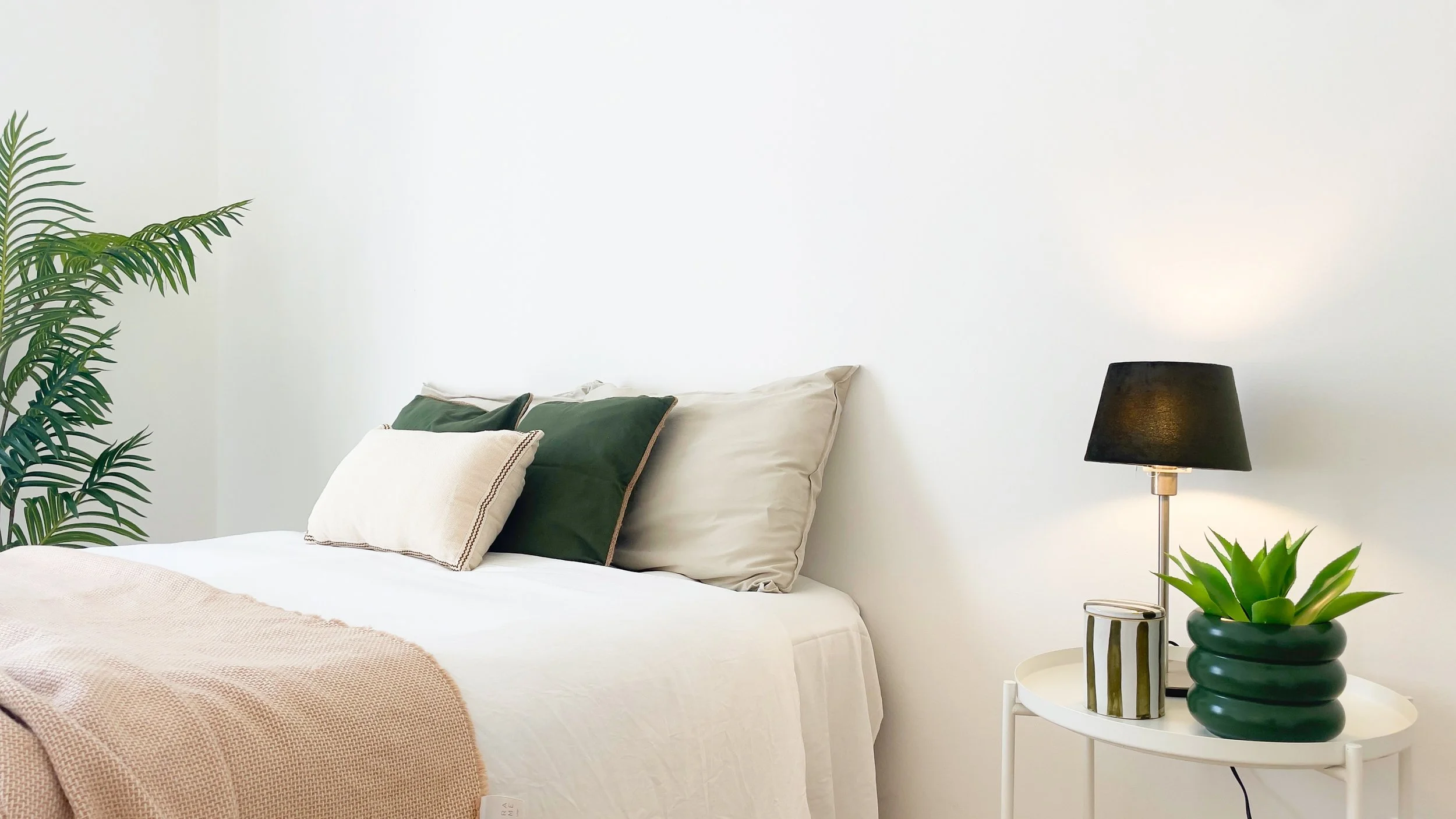 Camera da letto moderna con letto, cuscini verde e beige, lampada da tavolo nera e pianta in vaso su comodino bianco.