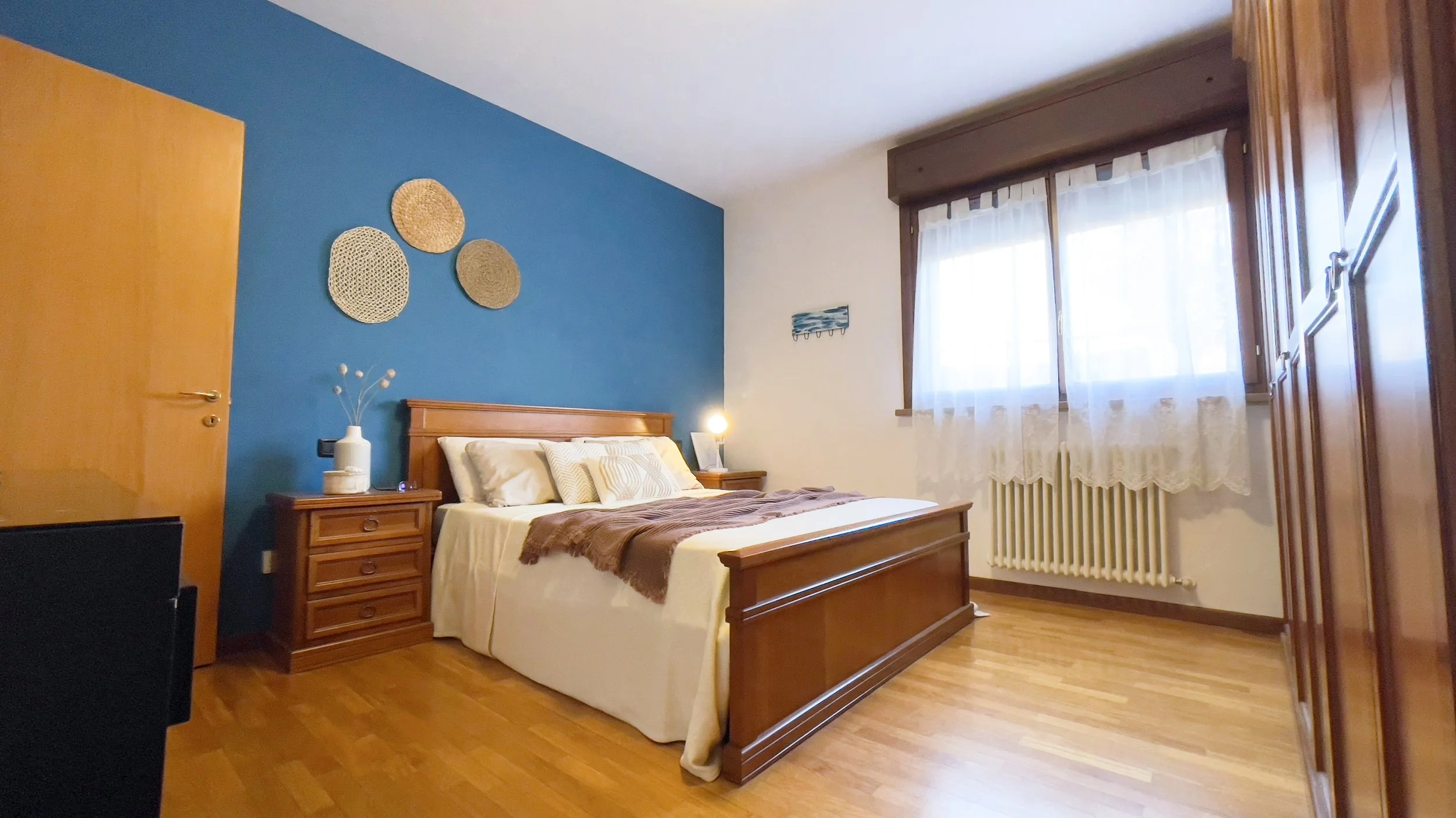 Camera da letto con pareti blu e bianche, letto matrimoniale in legno con coperte beige e marroni, comodini con lampade, finestre con tende bianche, pavimento in legno e decorazioni rotonde sulla parete.