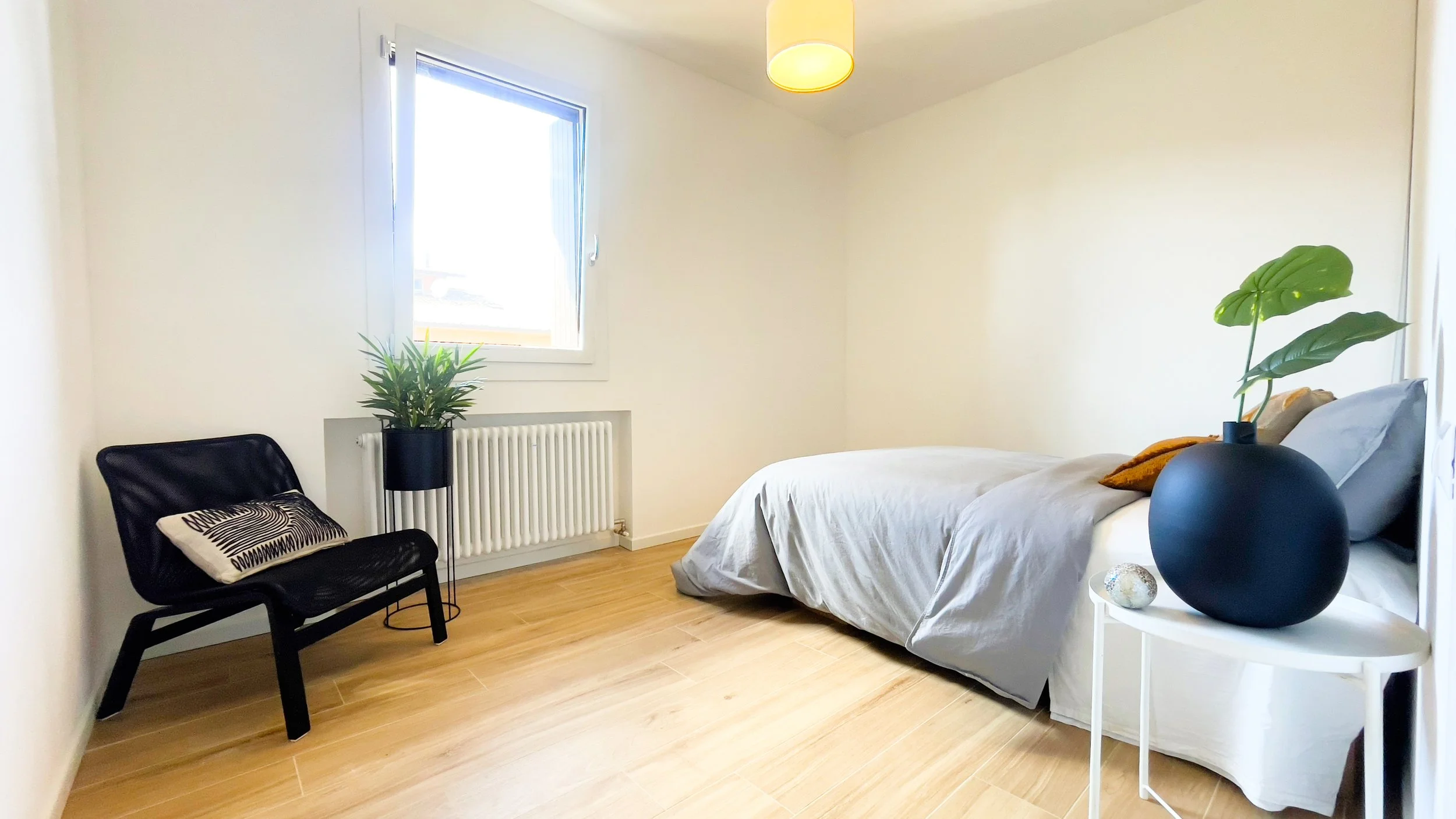 Camera da letto minimalista con letto grigio, sedia nera con cuscino decorativo, pianta su supporto, tavolino bianco con vaso e finestra con luce naturale.