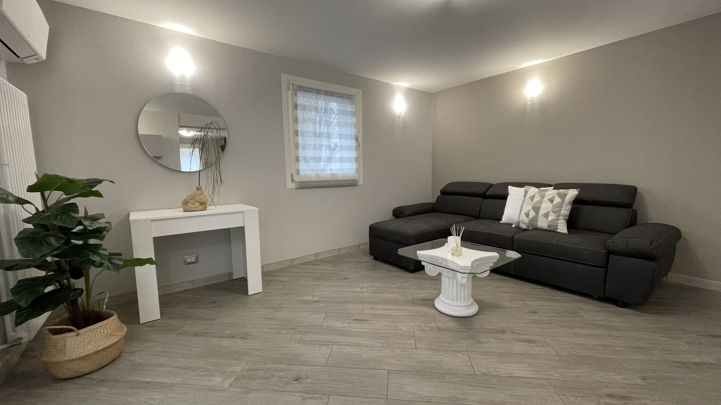 Soggiorno moderno con divano scuro, tavolino in vetro su piedistallo bianco, specchio rotondo sopra un tavolo bianco e pianta in vaso intrecciato.