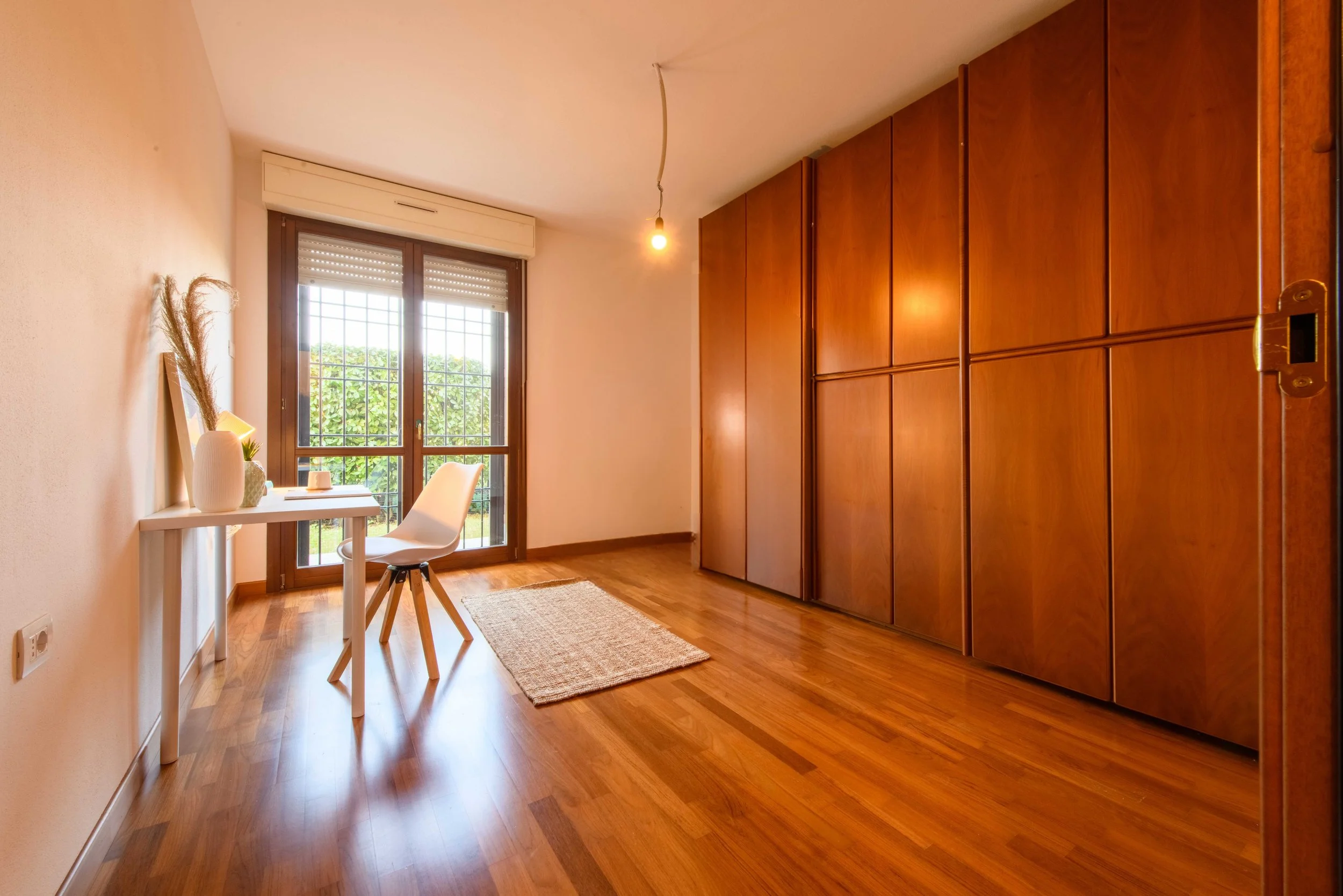Stanza luminosa con pavimento in legno, scrivania bianca con sedia e decorazioni, armadio in legno, finestra con tende abbassate, lampadina sospesa e tappeto beige.