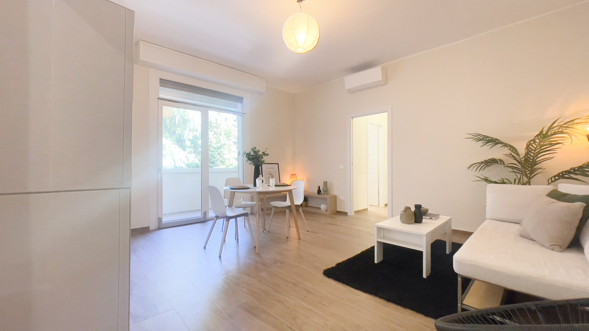 Soggiorno luminoso con arredamento moderno, tavolo da pranzo rotondo con sedie bianche, divano bianco, tappeto nero, pianta decorativa e lampada a sospensione. Porta finestra che conduce a un balcone.