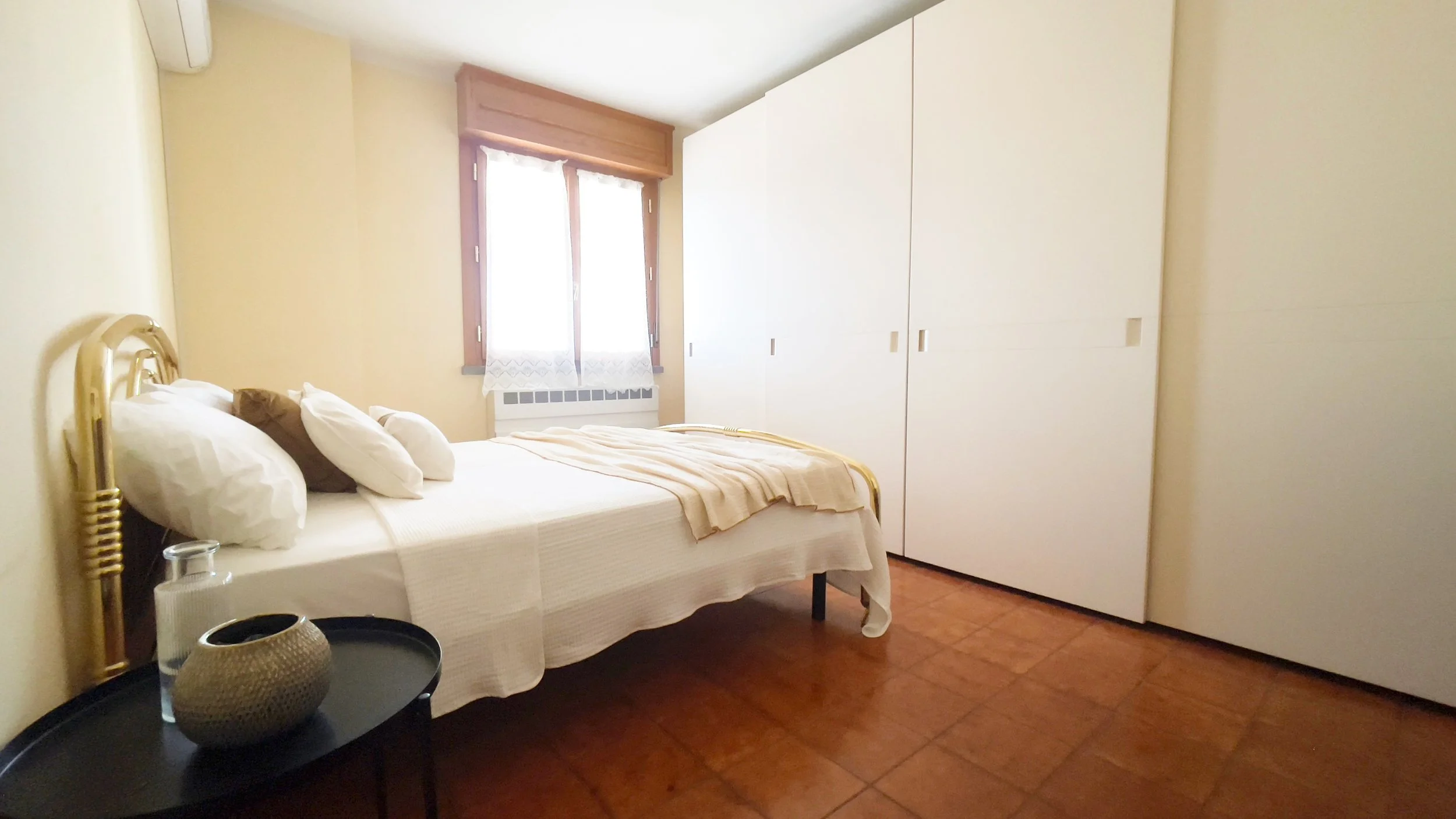 Camera da letto con letto matrimoniale, armadio bianco, finestra con tende, tavolino con oggetti decorativi.