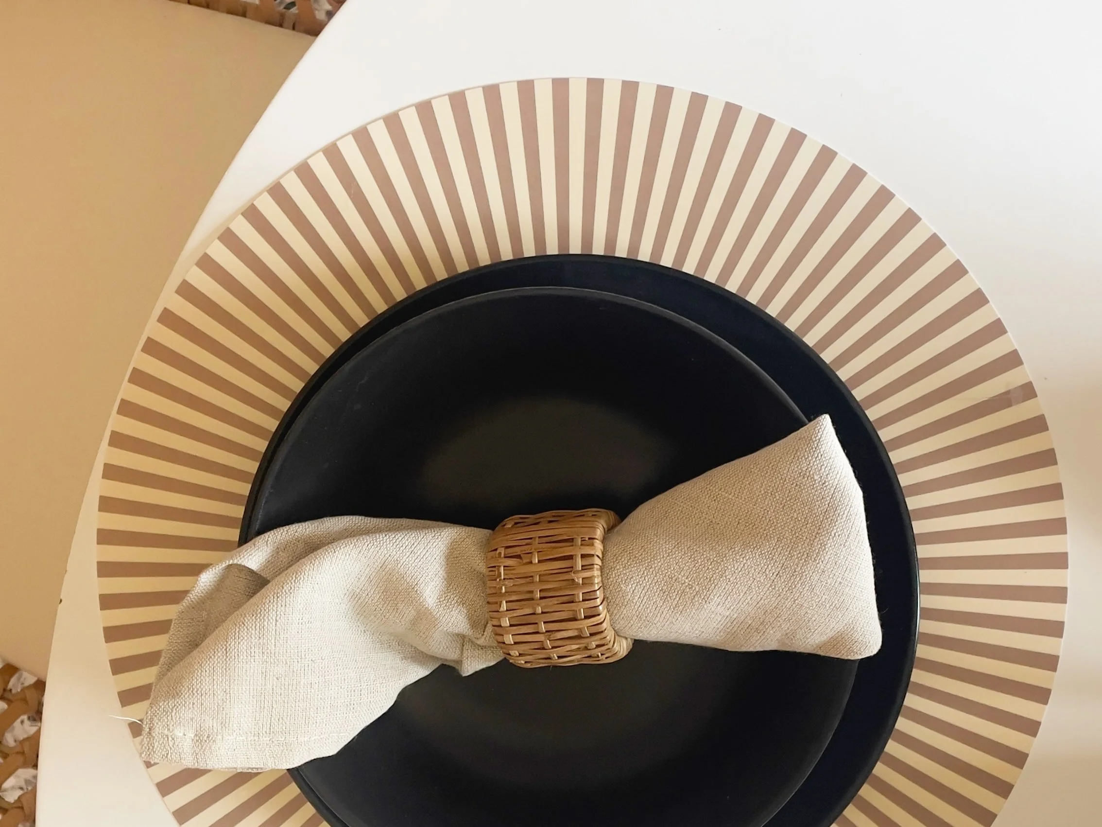 Immagine di un tavolo apparecchiato con un piatto nero sopra un sottopiatto a strisce marroni e beige. Al centro, un tovagliolo di stoffa chiaro avvolto in un portatovagliolo intrecciato.